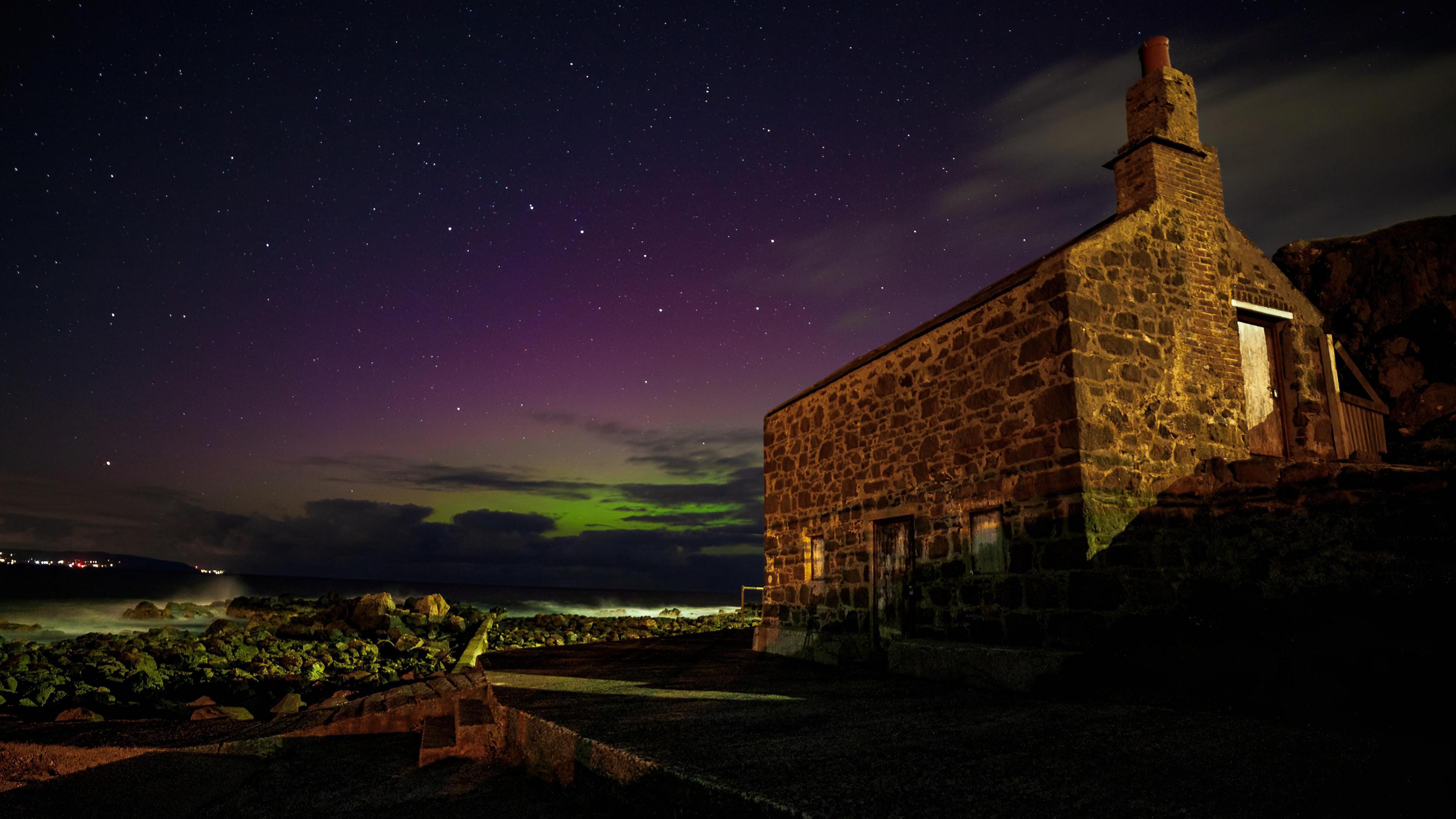 A grey stone cottage sits in the foreground of a green-purple glow form the sky behind and above it