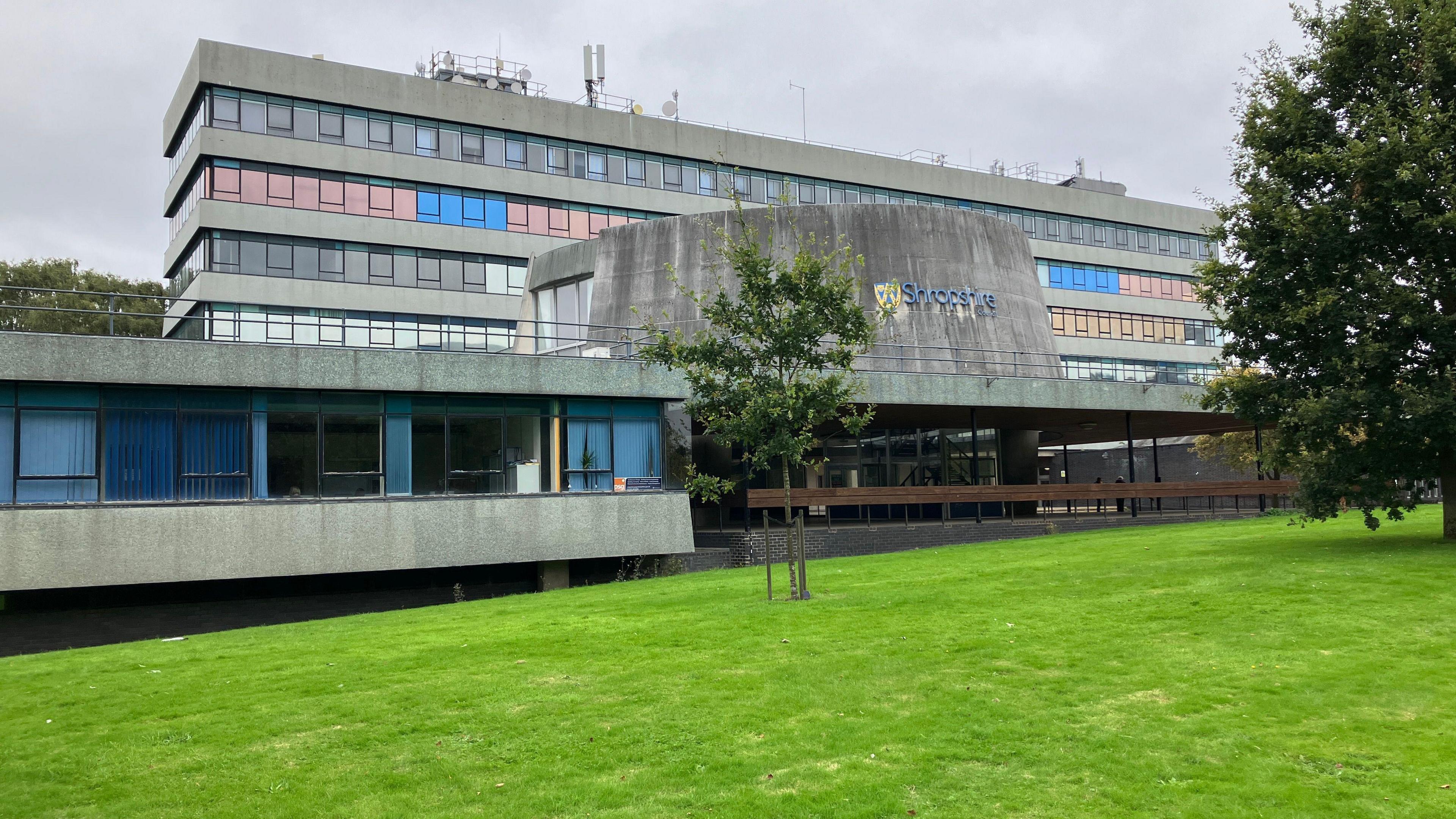 Large building that is made of grey concrete with rows of dozens of windows. There is a large funnel-shaped structure above an entryway, with a sign on it that reads "Shropshire Council".