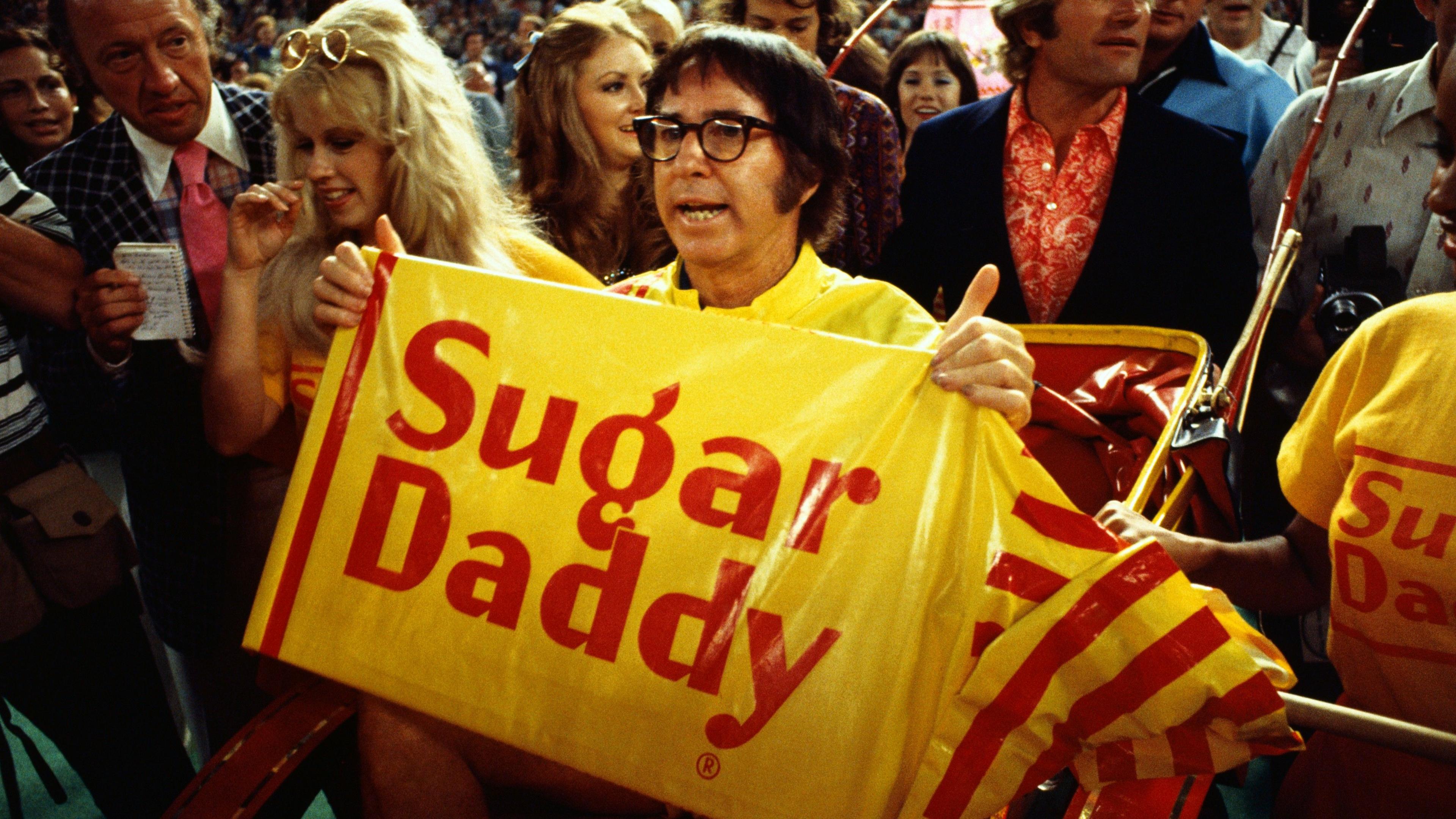 Bobby Riggs holding a "Sugar Daddy" sign while being carried to the court by young women before the Battle of the Sexes match