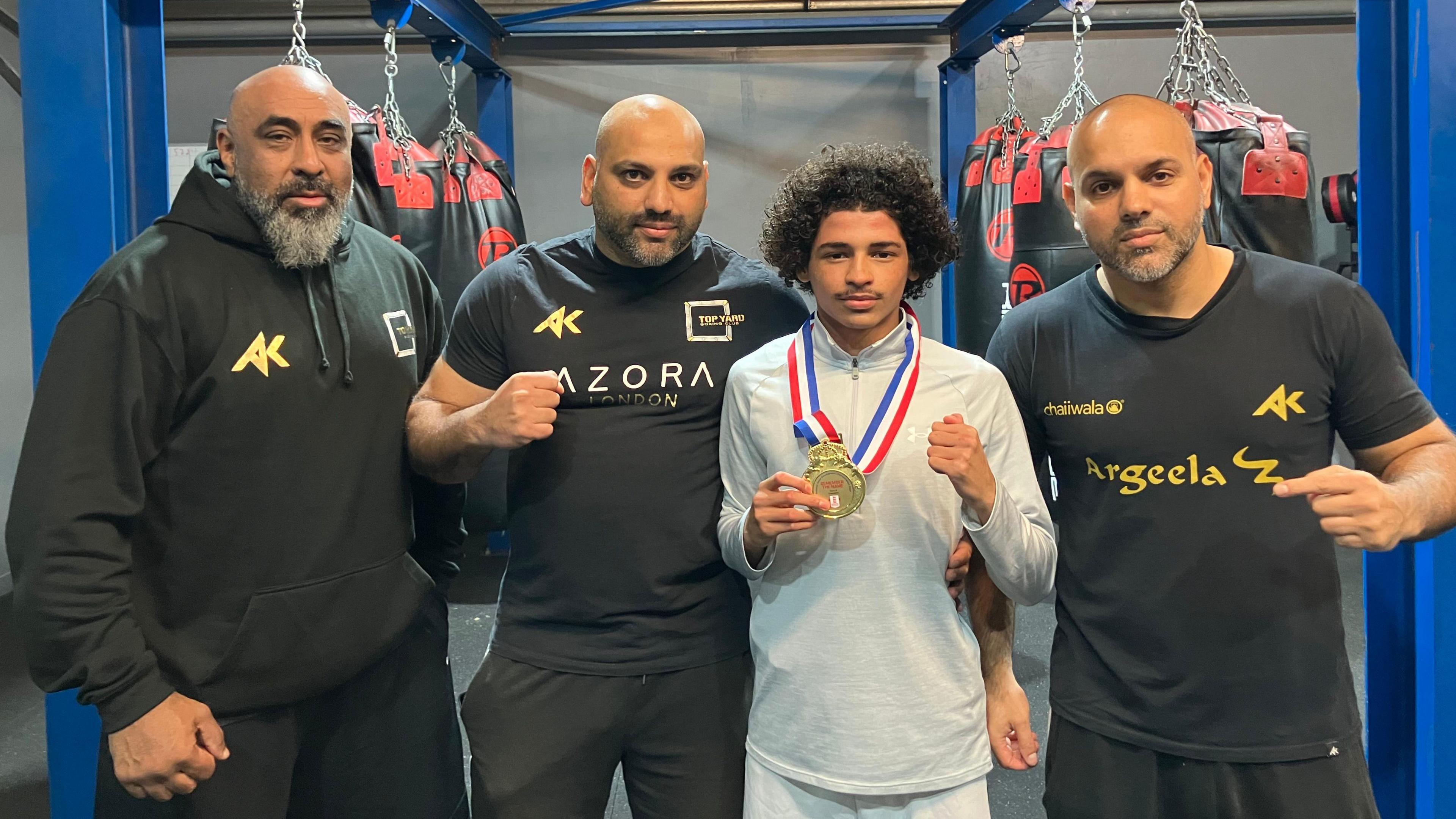 Shujah wearing a black Hoodie, next to Hamad wearing a black T-shirt, standing next to Ismael wearing a grey track suit and a gold medal around his neck and Bilal wearing a black t-shirt - fist pumping, smiling for camera 