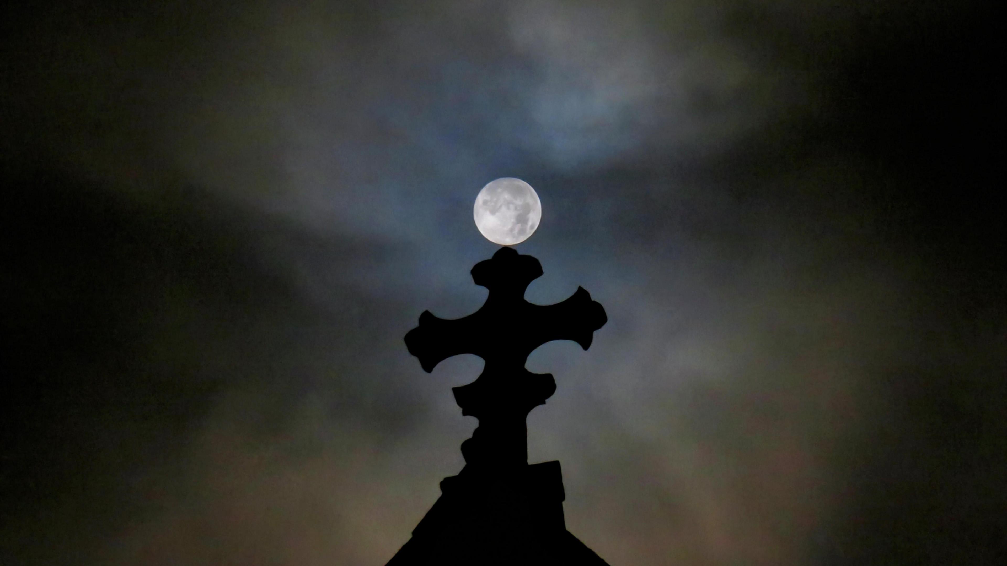 A nighttime shot of the top of an apparent church building in shadow with the moon in the sky above. The sky is mainly black but there is a slight white glow around the moon. The top of the church is in the shape of a cross which is in darkness.