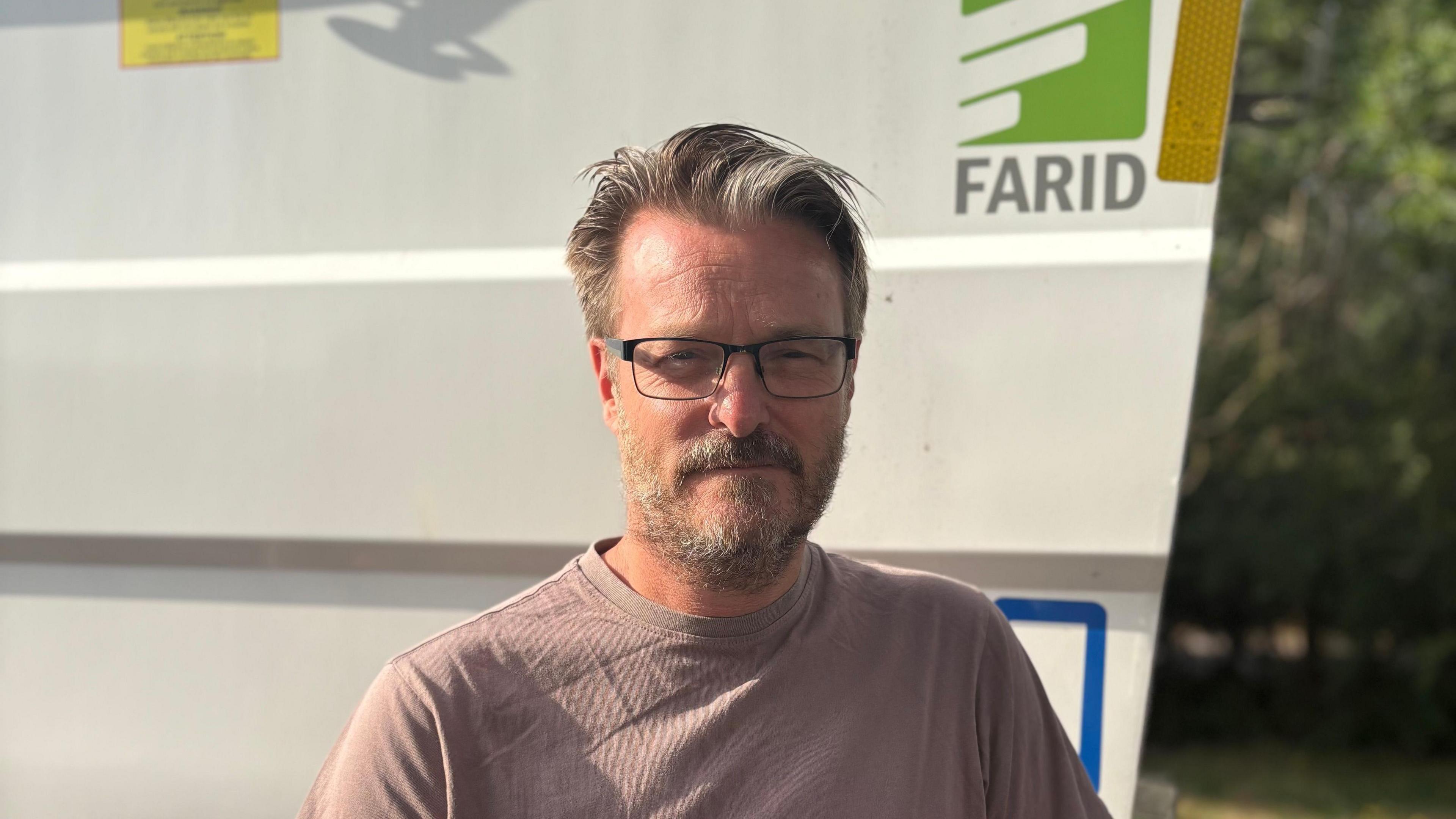 A man wearing glasses and with a greying beard smiles at the camera while standing in front of a recycling truck bearing the word "Farid".