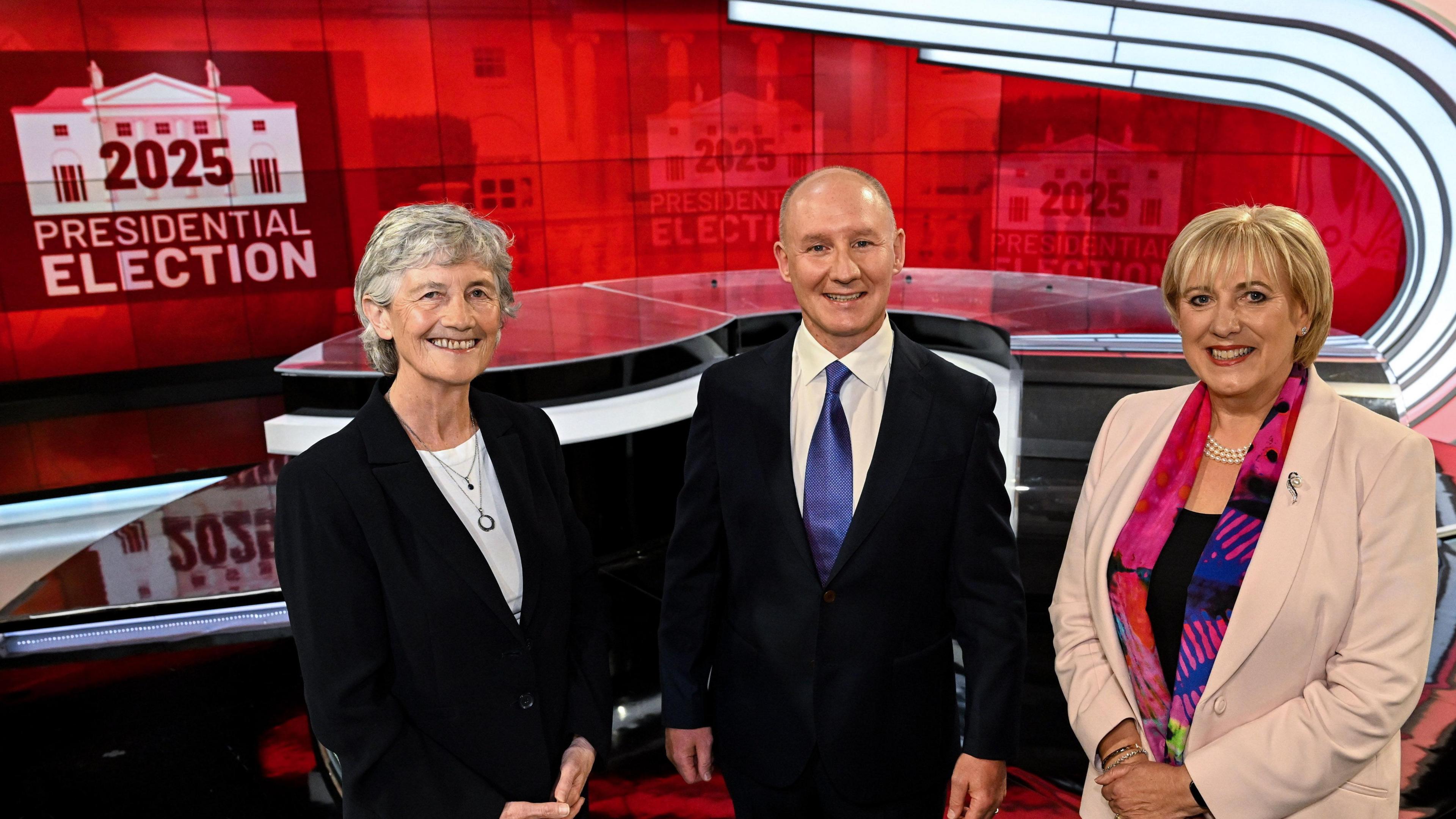 Catherine Connolly, Jim Gavin and Heather Humphreys stand in a TV studio smiling at the camera. Behind them is a large red screen displaying a 2025 Presidential Election logo. Catherine Connolly on the left is an older woman, with short grey hair, a black suit jacket and a white shirt. In the middle is Jim Gavin, who is a bald man and is wearing a black suit jacket, a white collared shirt and a blue tie. On the right is Heather Humphreys, who is an older woman with short blonde hair. She is wearing a ligh coloured suit jacket, a colourful scarf and a black top.