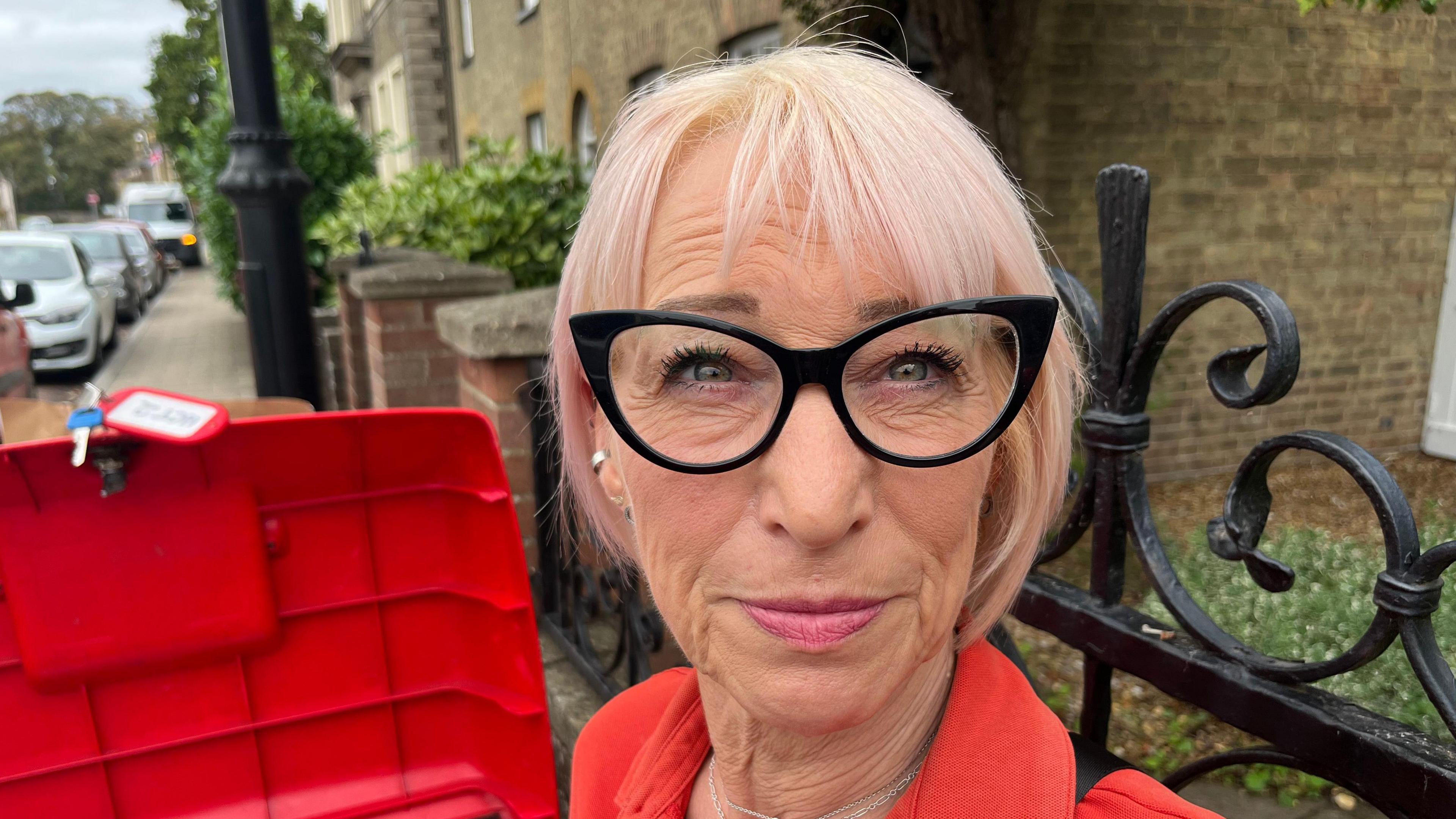 Sarah Dunsmore has short blonde hair in a bob style. She has black framed glasses. She is wearing a red top and behind her is a row of houses. She has two thin silver chains around her neck.