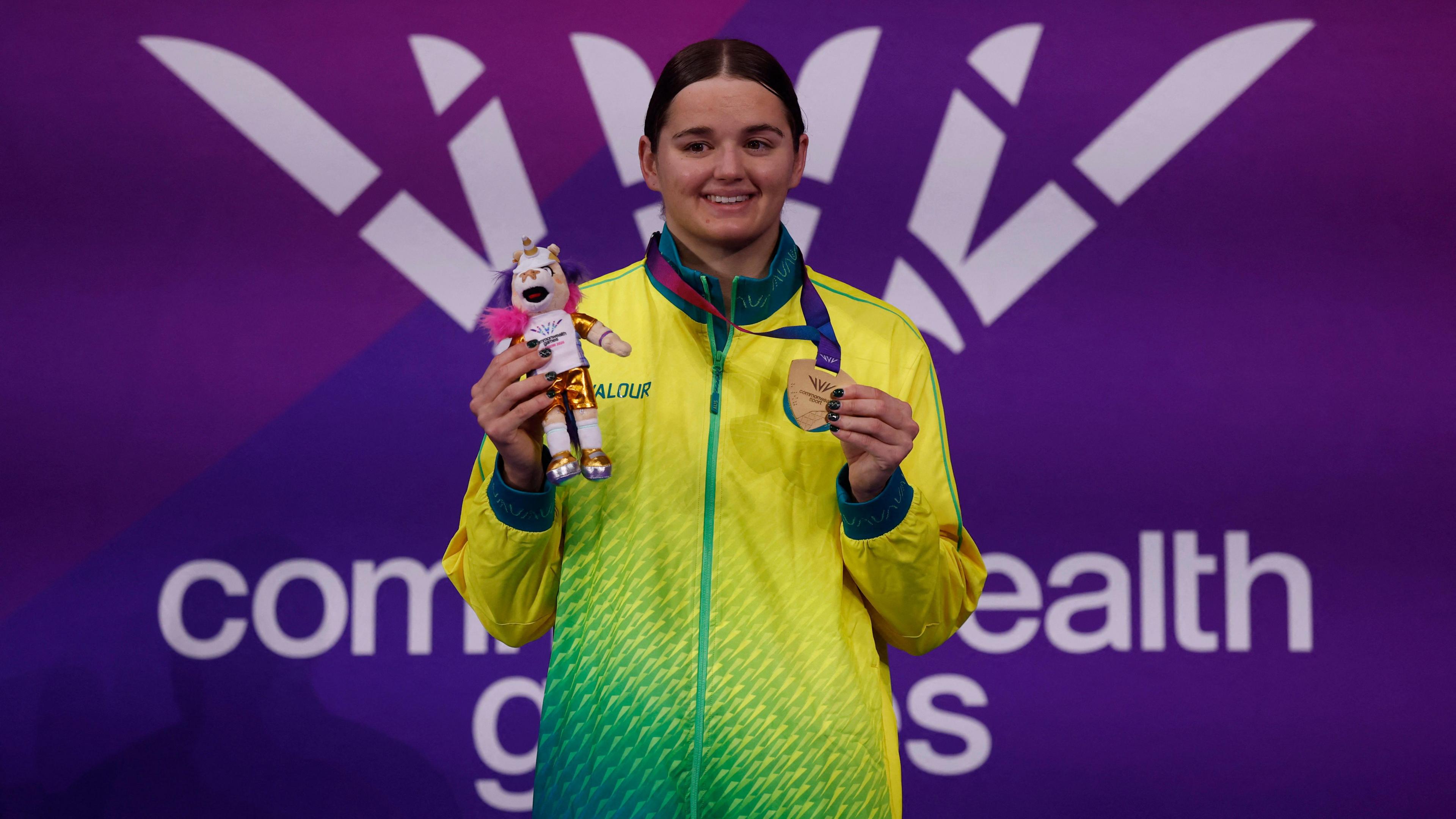 Iona Anderson - a swimmer wearing a green and yellow tracksuit top, holds a gold medal. It is round her neck and she is holding it in one hand. In her other hand is a stuffed toy of Finnie - the unicorn mascot of the Commonwealth Games.