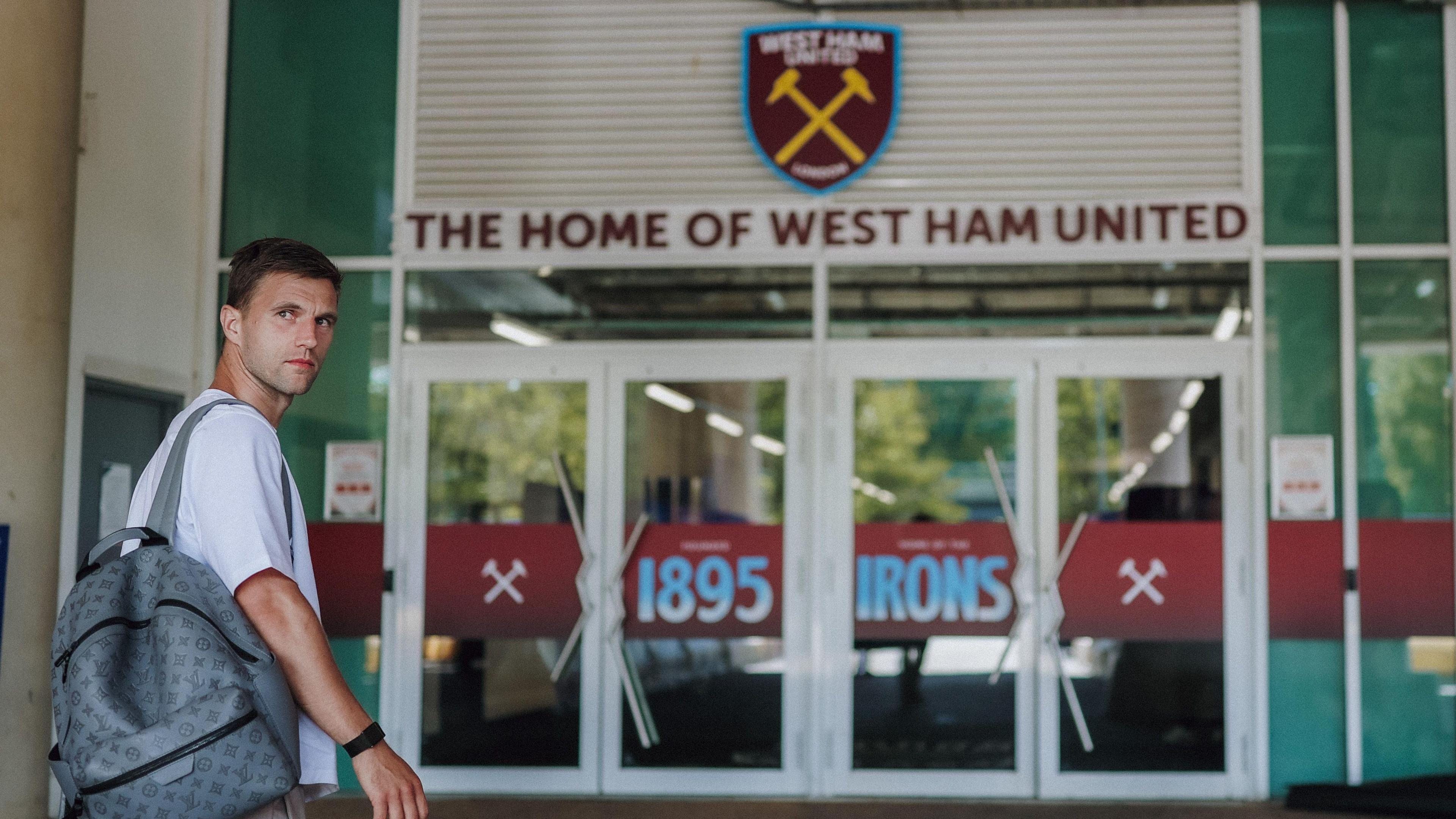 Joel Veltman, wearing a grey backpack and a white T-shirt, looks back at the camera in front of the entrance to London Stadium with the West Ham United club crest above the doors