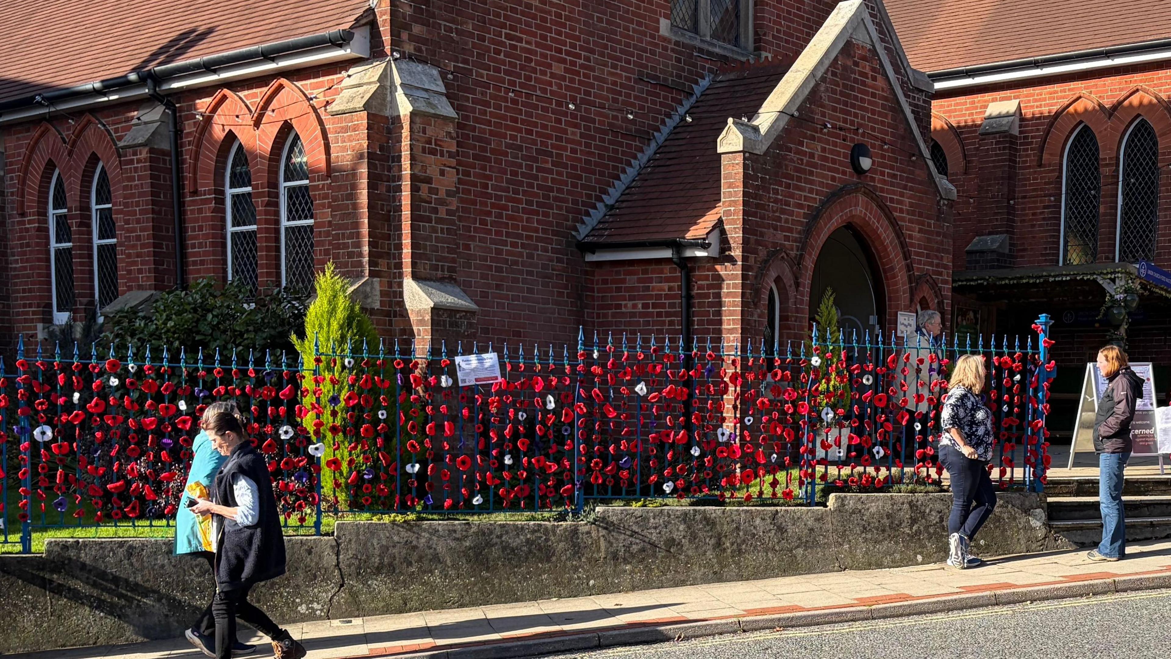 Red, purple and white poppies are attached to metal gates outside a church. There are several people walking pass the church.