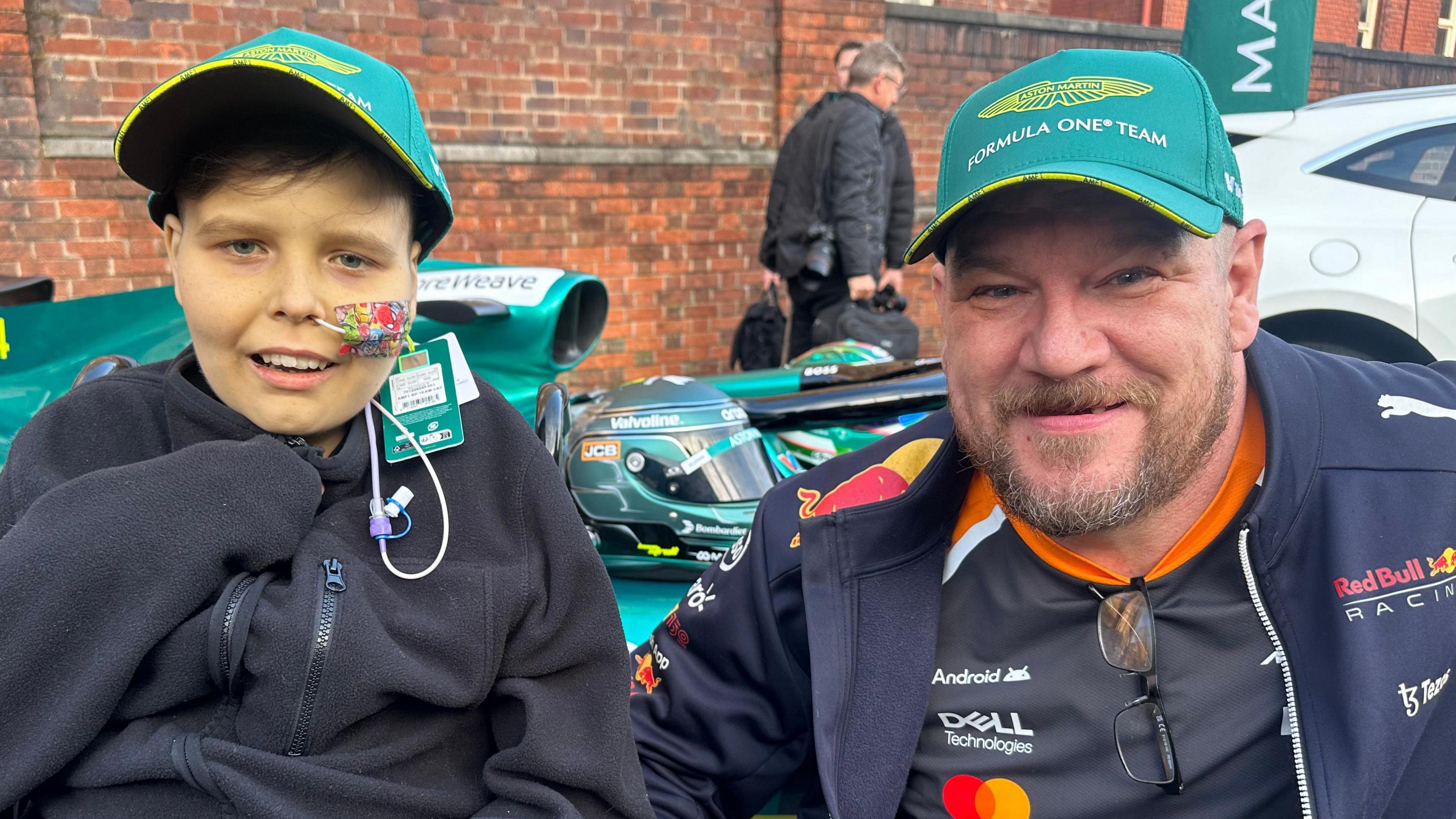 A man and son, both wearing green F1 hats, sat in front of a green F1 replica car. The boy is wearing a black fleece and has a tube going into his nose. The man is wearing an F1 jacket.