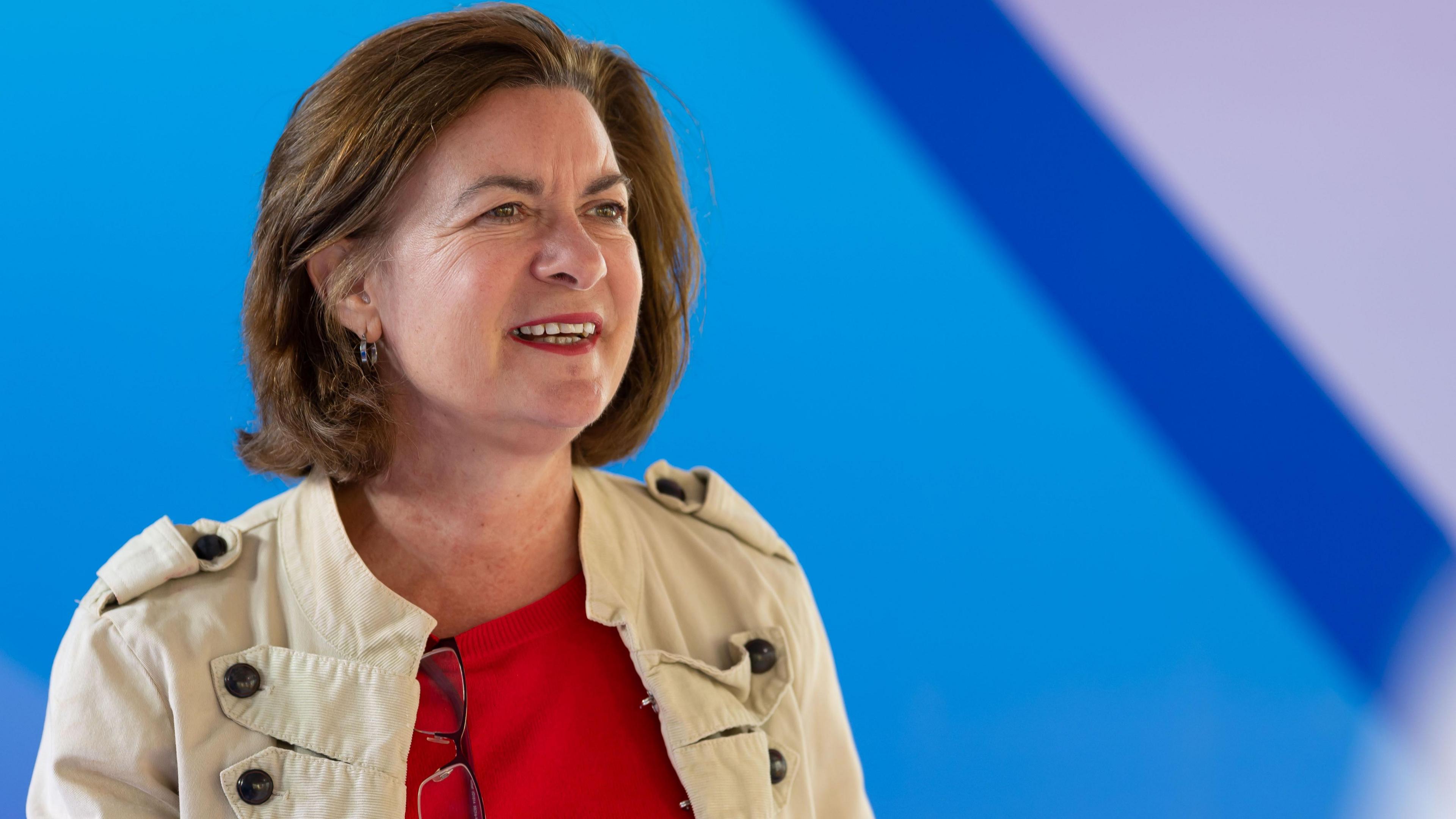 Eluned Morgan - A woman with brown hair looking to the left of the camera. She is wearing a red top and a beige jacket. There is a blue background 
