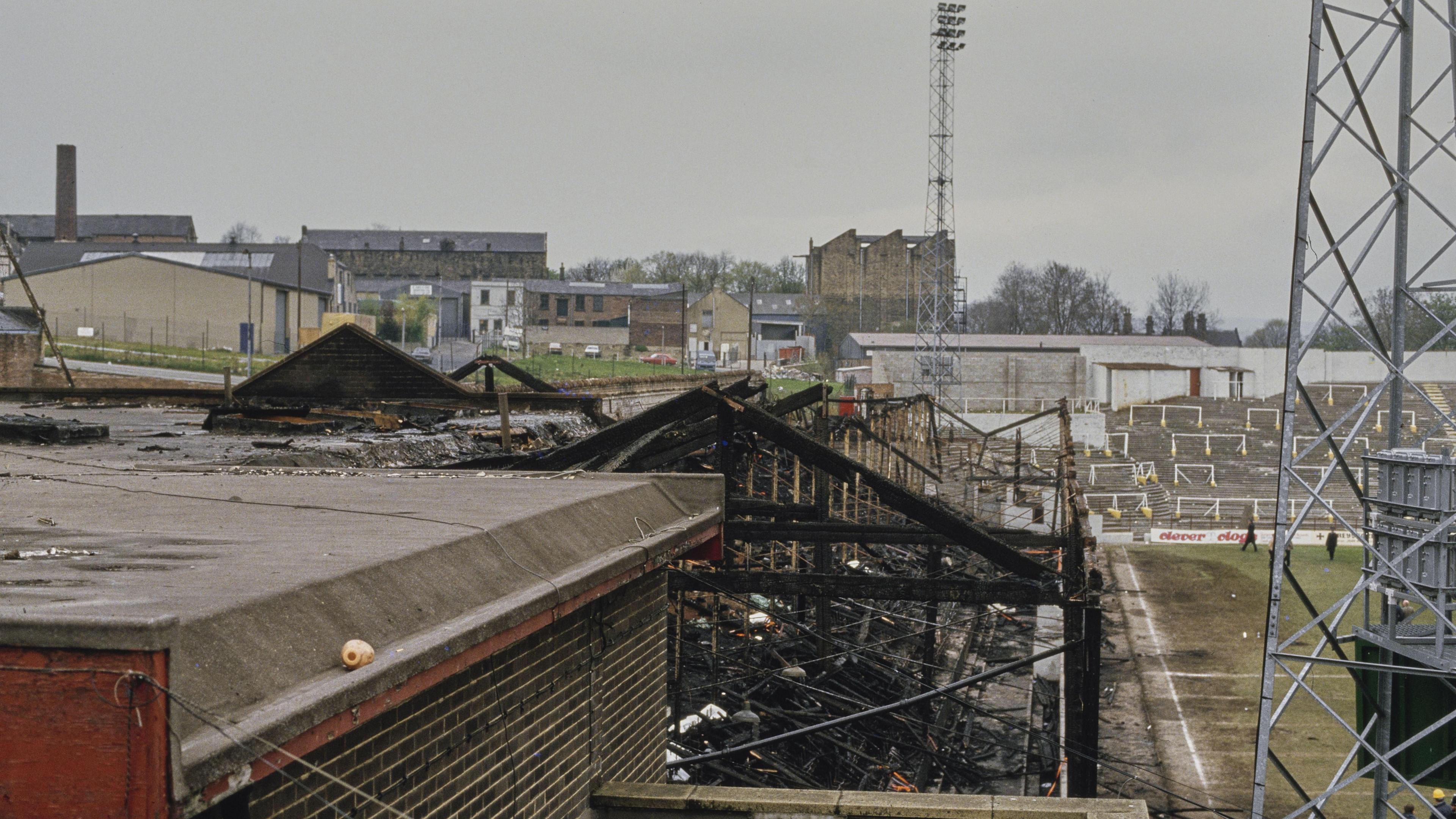 The fire-damaged skeleton of a football stand at Bradford City's Valley Parade ground, pictured in 1985. Another terraced stand, undamaged, can be seen in the distance, with an electric pylon in the foreground.