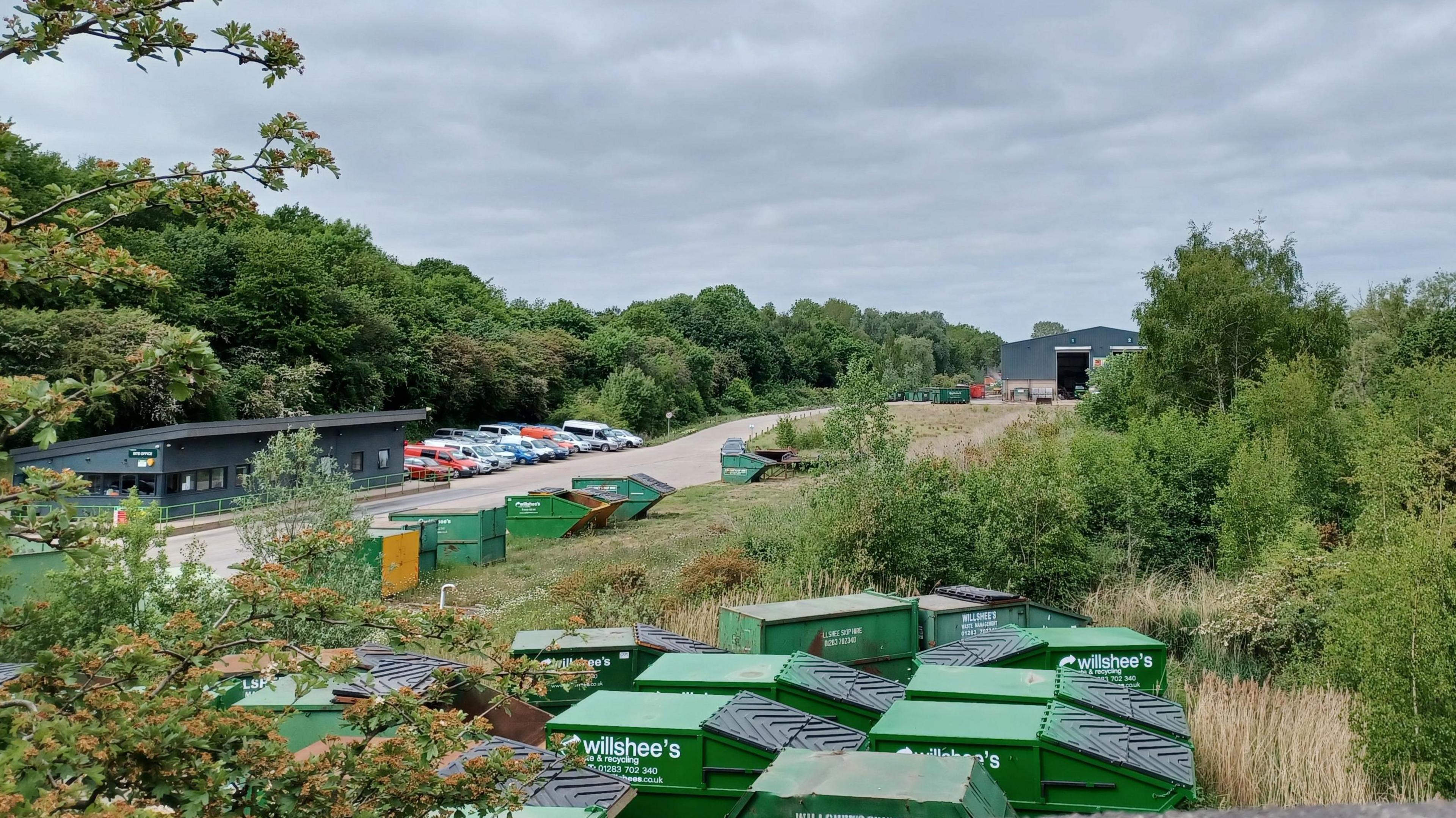 A cloudy sky overlooks a recycling center filled with green skip bins, surrounded by trees and parked cars in the distance