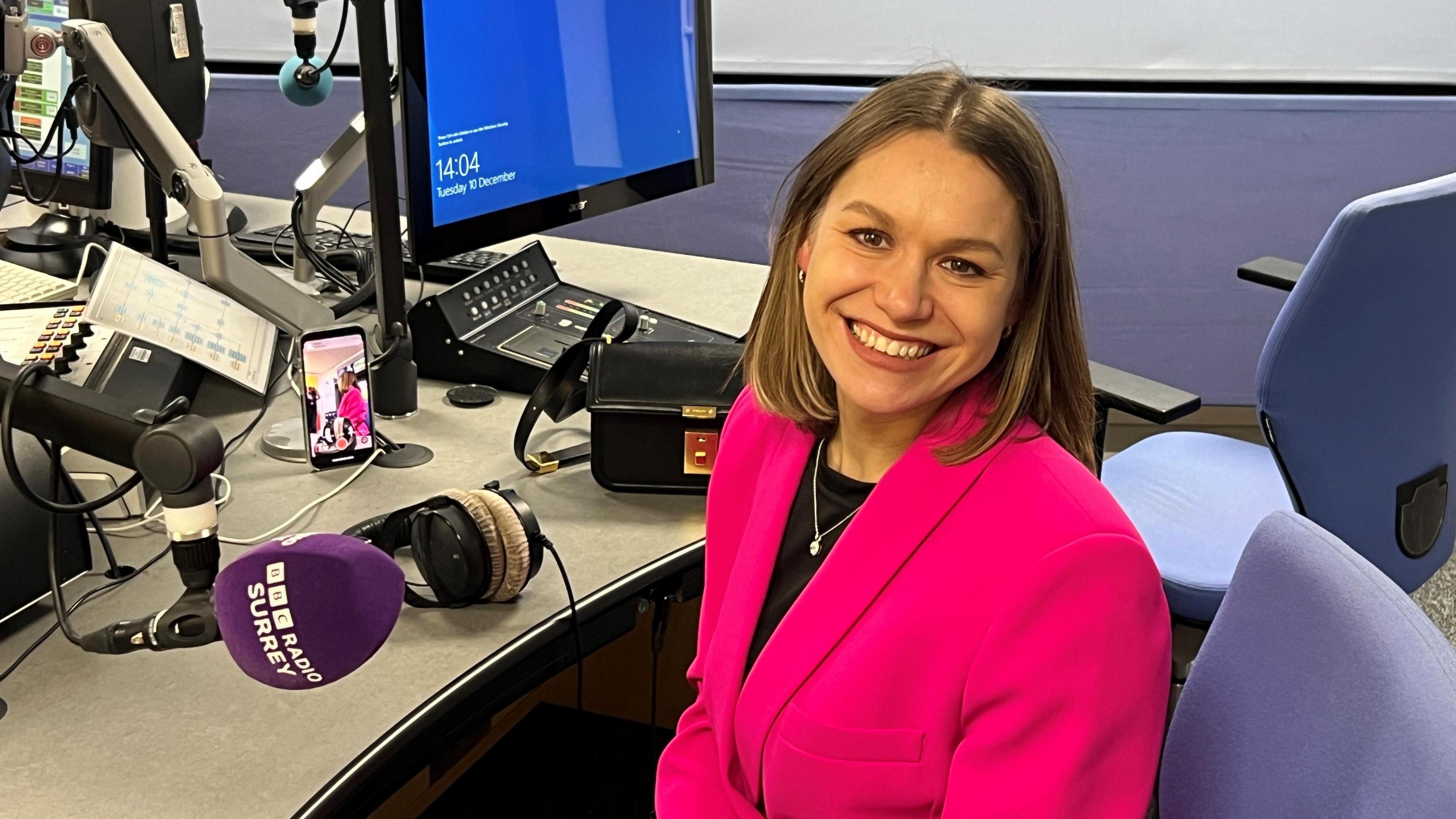 Young woman with brown hair and a bright pink jacket smiling and looking at the camera in a radio studio and in front of a purple microphone with BBC Radio Surrey logo on it. 