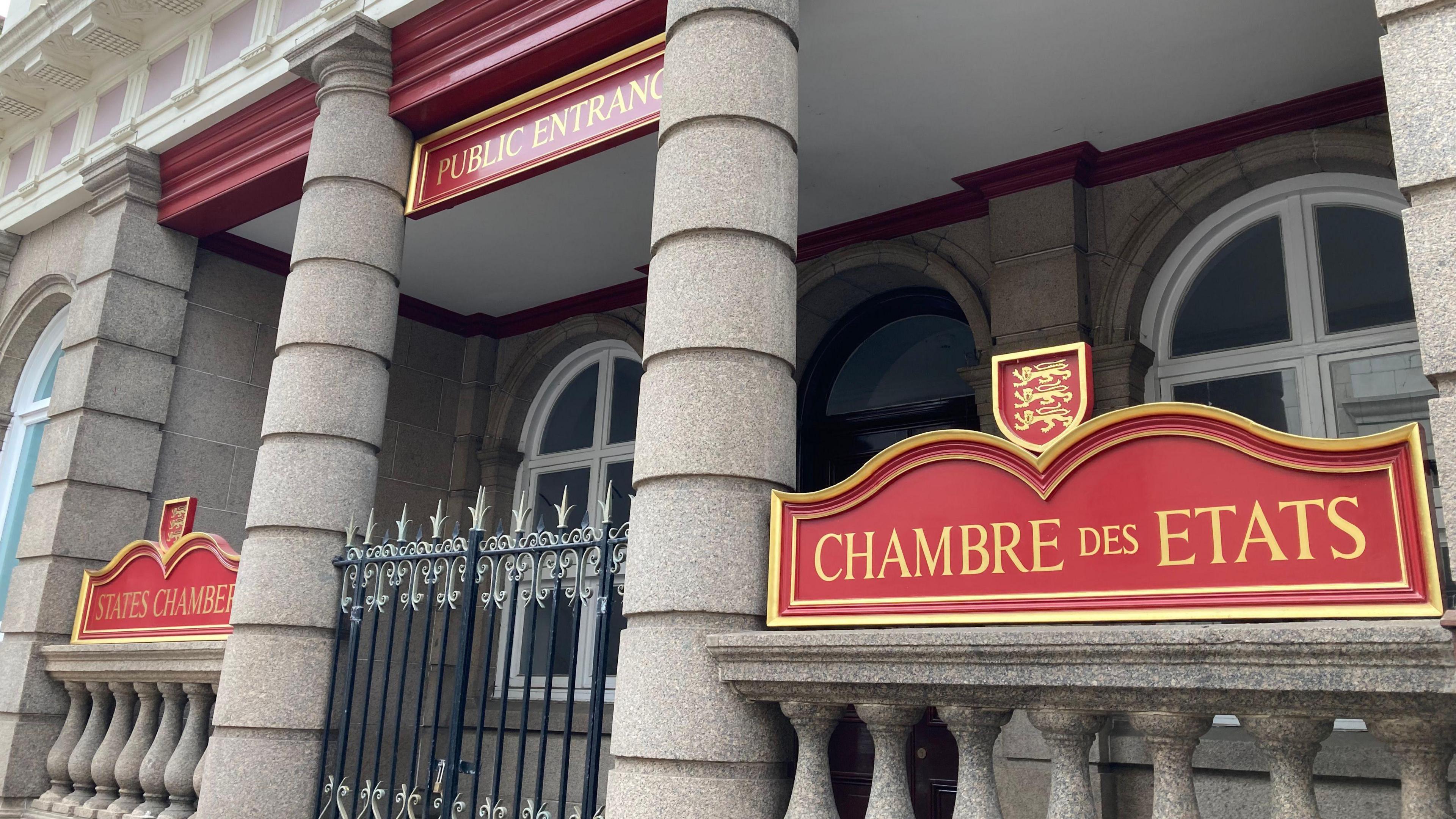 Entrance to a government building with three red and gold signs reading 'PUBLIC ENTRANCE,' 'STATES CHAMBER,' and 'CHAMBRE DES ETATS.' The building features large stone columns and arched windows, suggesting an official or legislative setting.