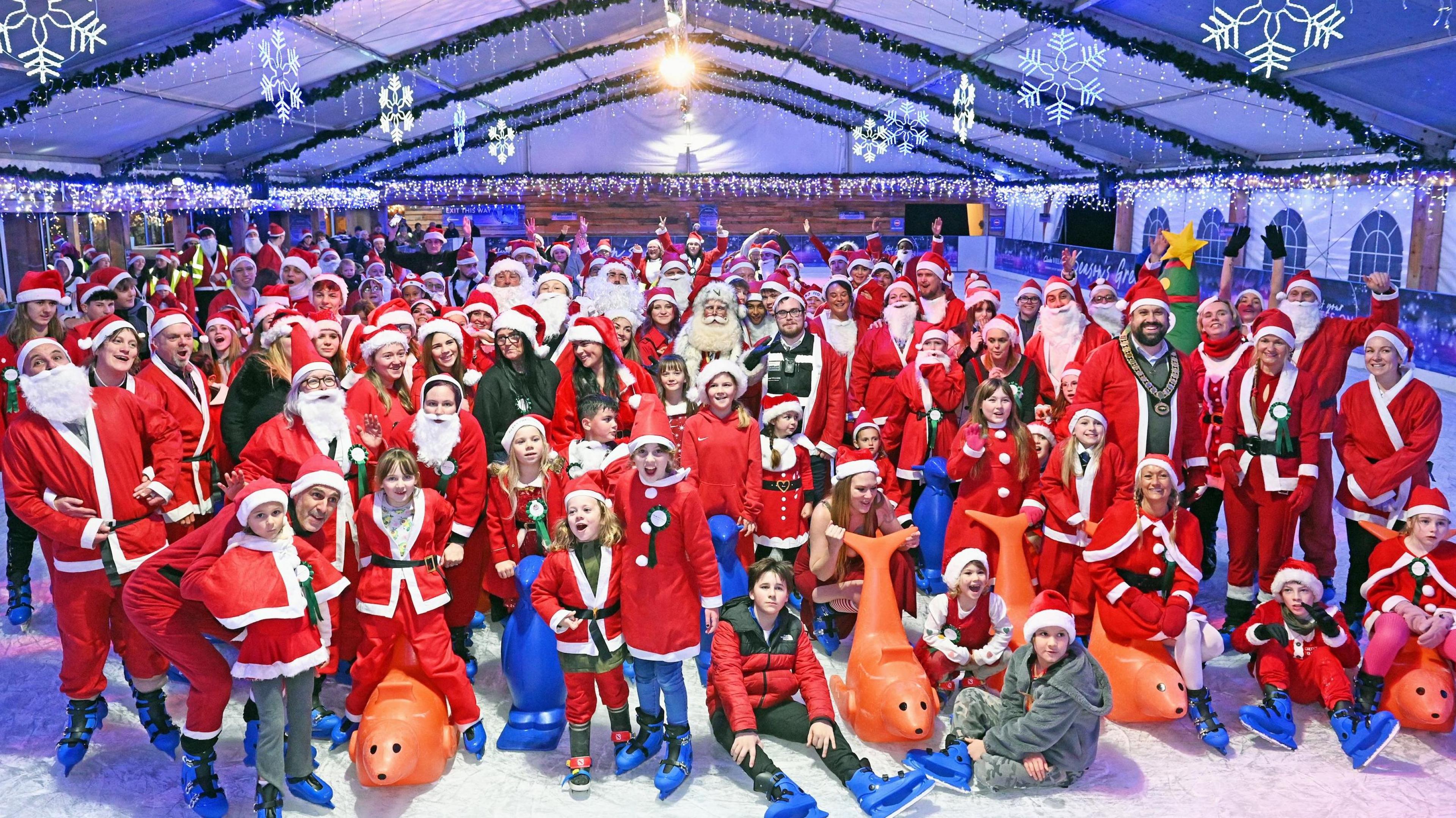 People on ice skating rink wearing red Santa outfits and blue skates. There is a range of ages with a man dressed as Santa in the middle.