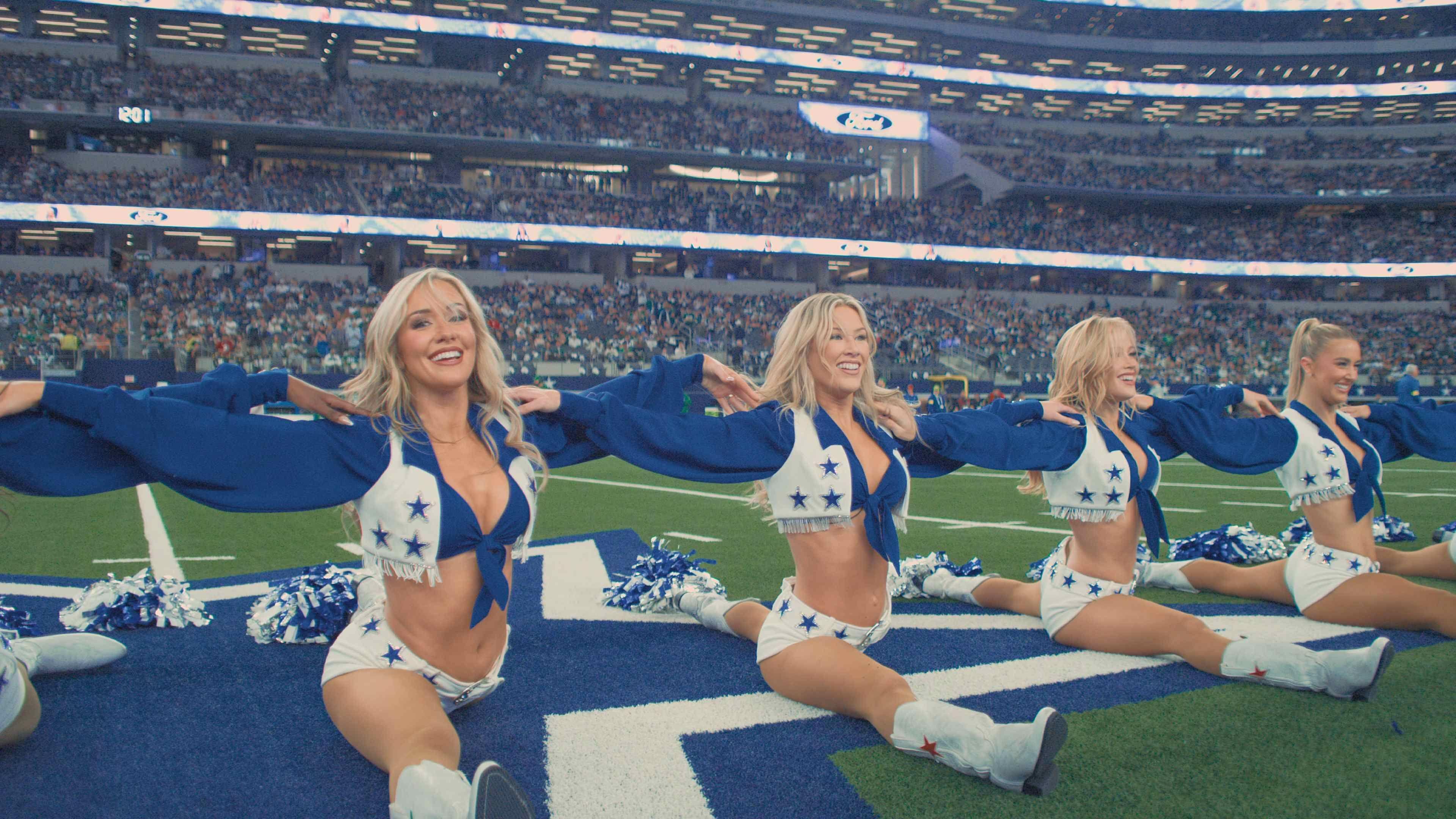 The Dallas Cowboys Cheerleaders in formation on the football field with crowds behind them. Four blonde cheerleaders look straight ahead with their legs flat, performing the splits.