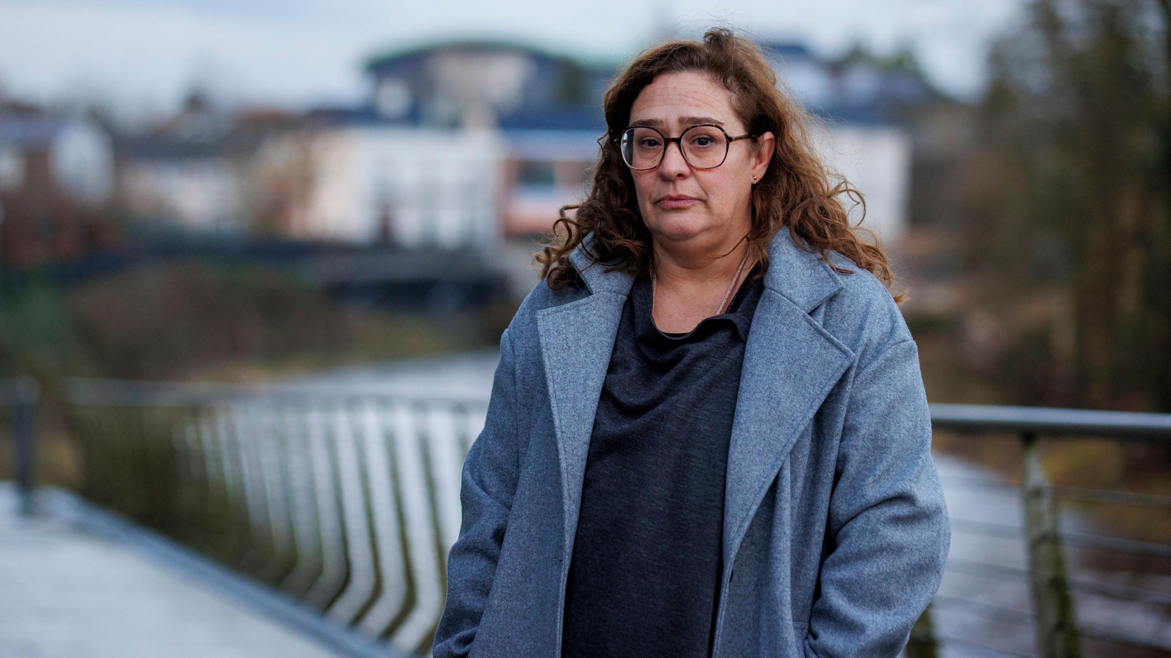 Paloma Abad Ramos looks at the camera. She has long brown curly hair, half of it tied back. She has large brown-rimmed glasses on and is wearing a grey coat, with a handbag strap visible over one shoulder.