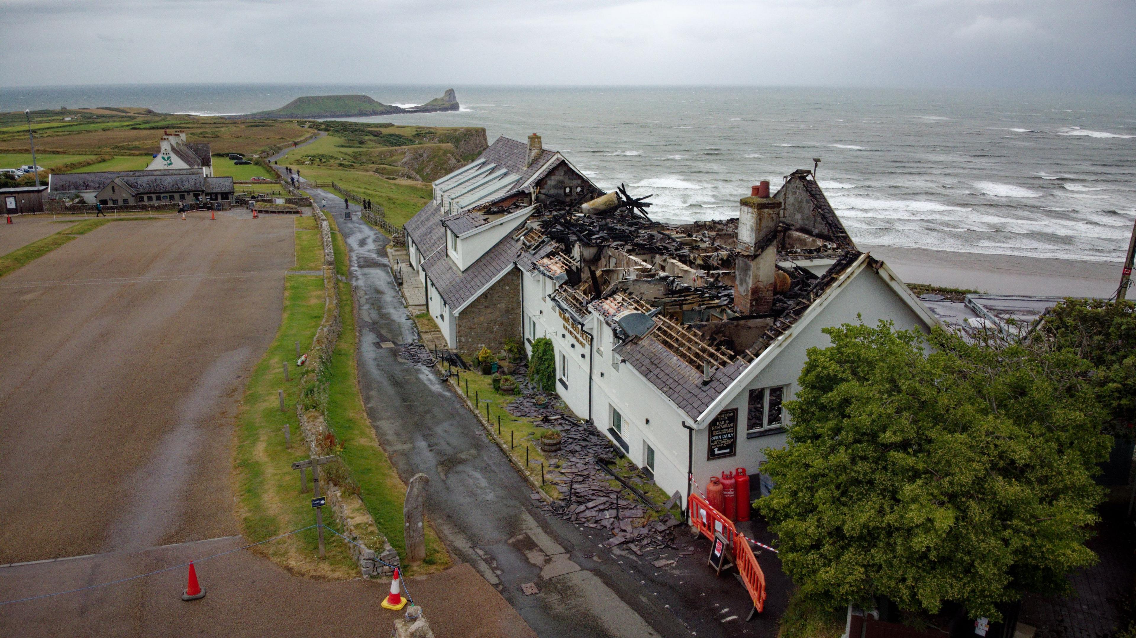 A wide image of the damage the fire has caused to The Worm's Head Hotel. The roof of the hotel has completely collapsed. Behind the hotel is a choppy sea and in front of it is a large empty car park. The picture has been taken on a cloudy day.