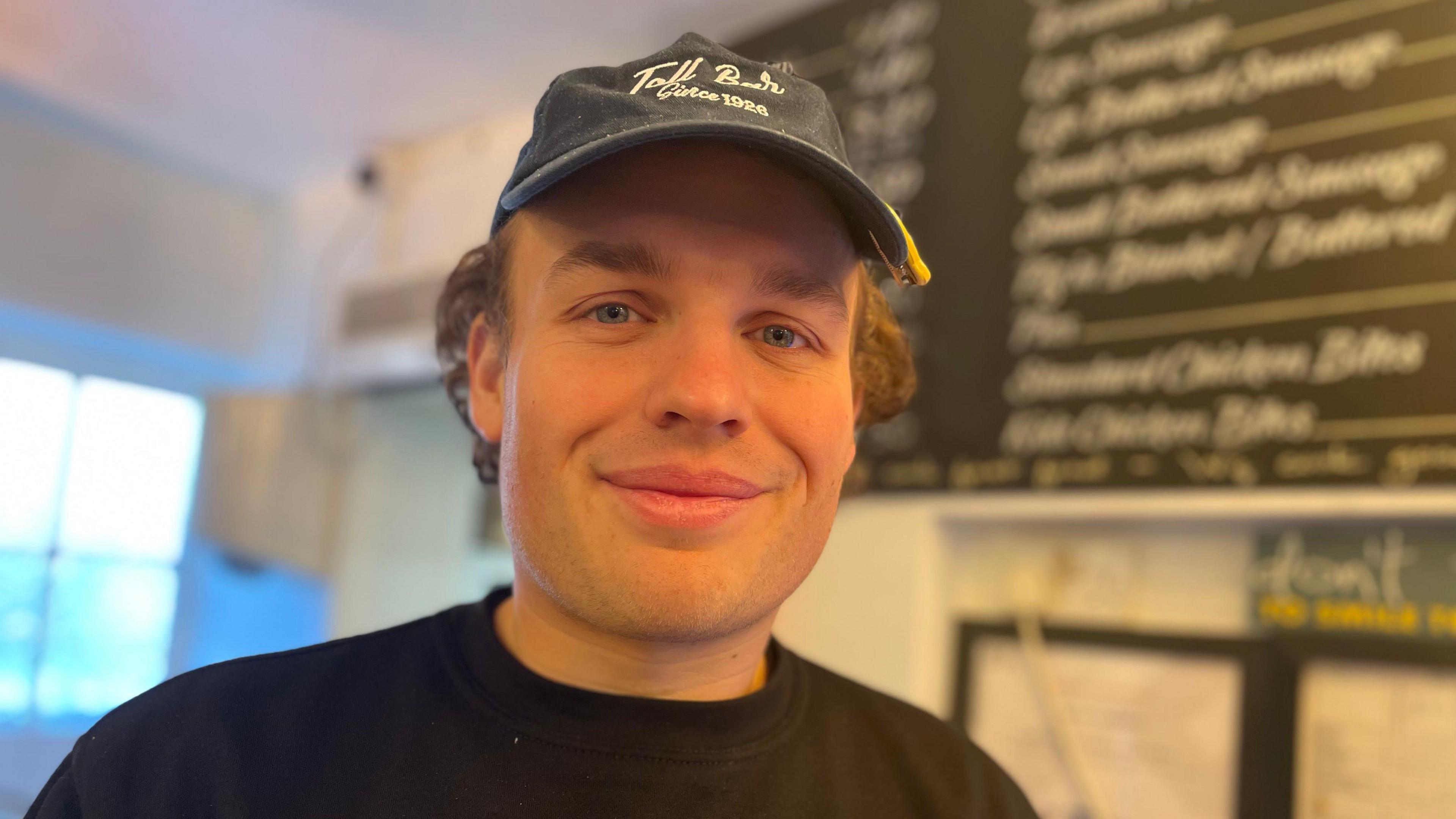 Young man in a black top and cap in front of a menu board