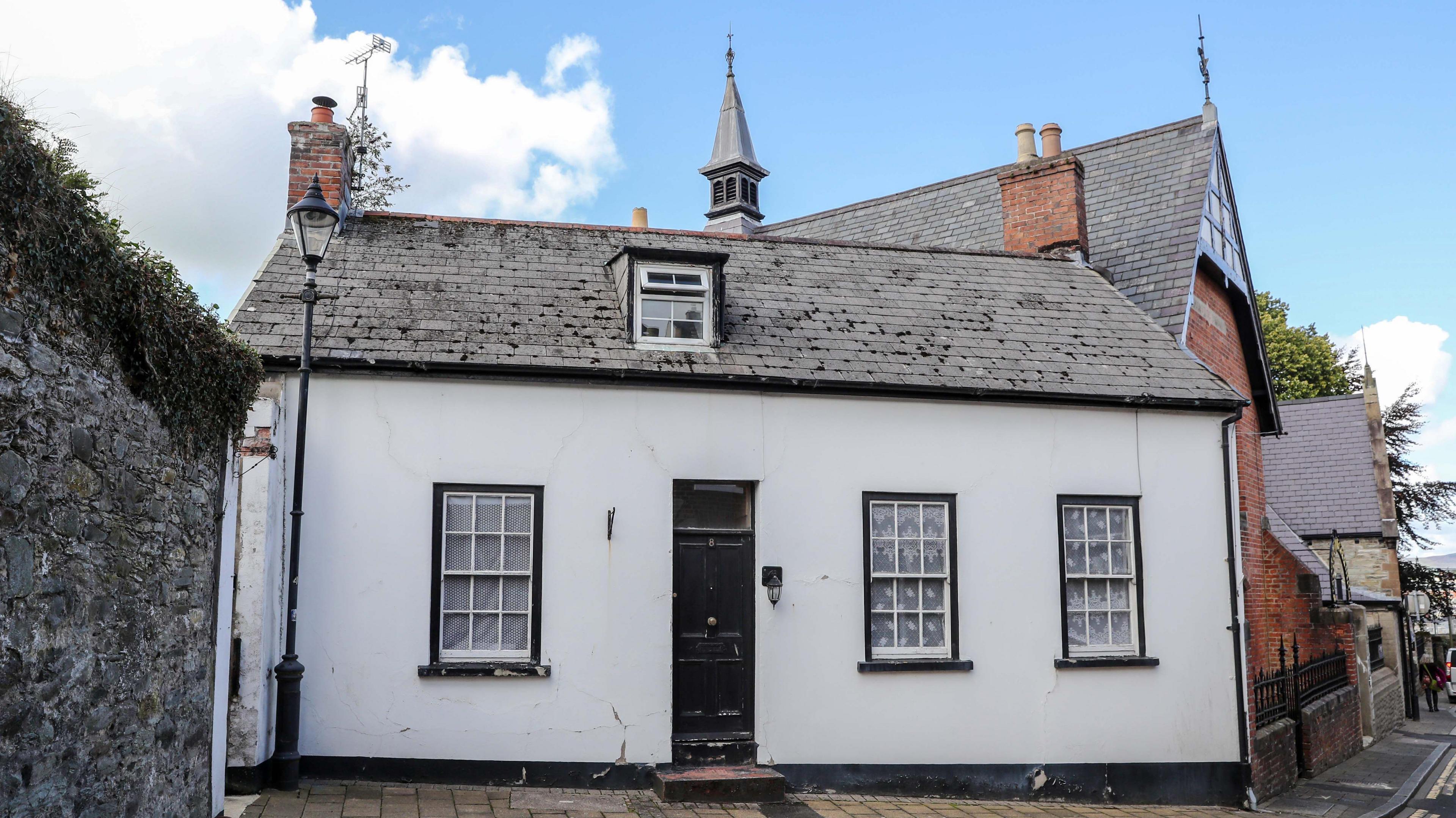 A cottage stands at the bottom of Palace Street in Londonderry. It is painted white on its exterior, on which there are three long windows and a door. A church building is in the background.
