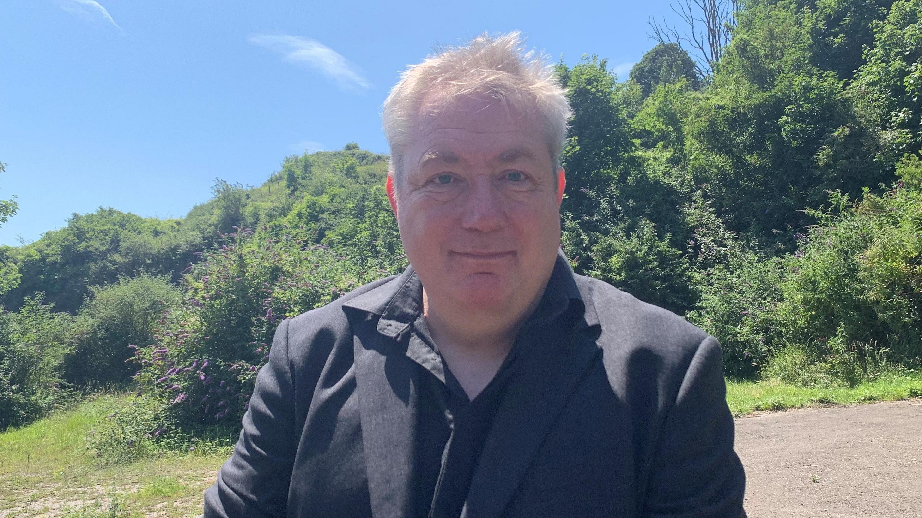 A middle aged man with grey hair wearing a black shirt and blazer is smiling. He is standing outside in front of some bushes on a sunny day.