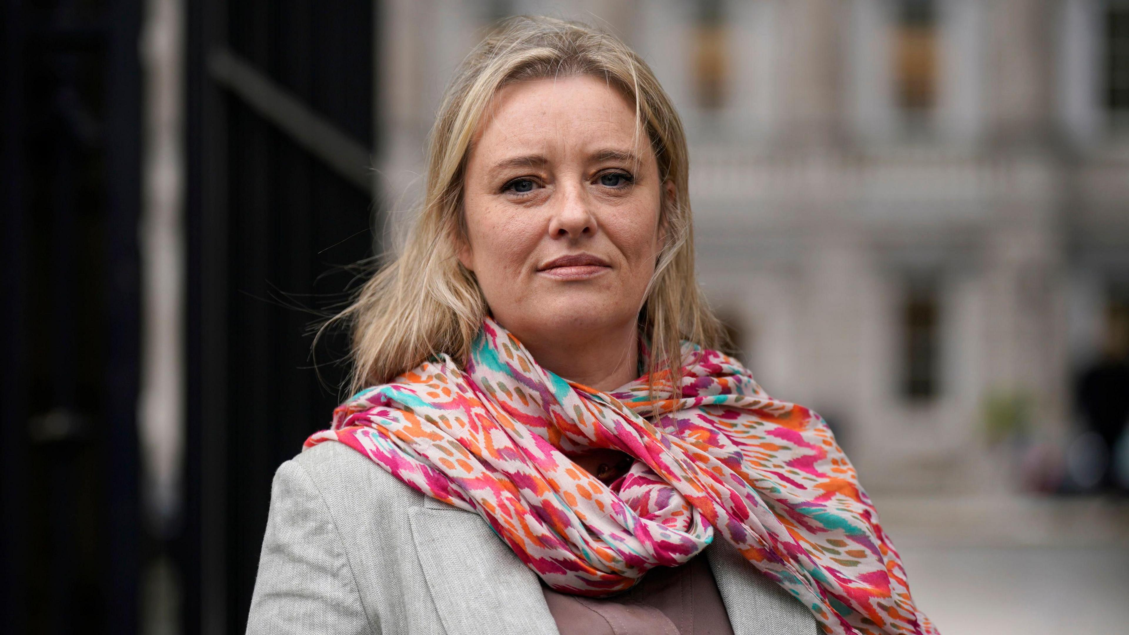 Máiría Cahill stands in front of the camera. She has blonde hair, a grey jacket and a colourful scarf. Her expression is neutral.