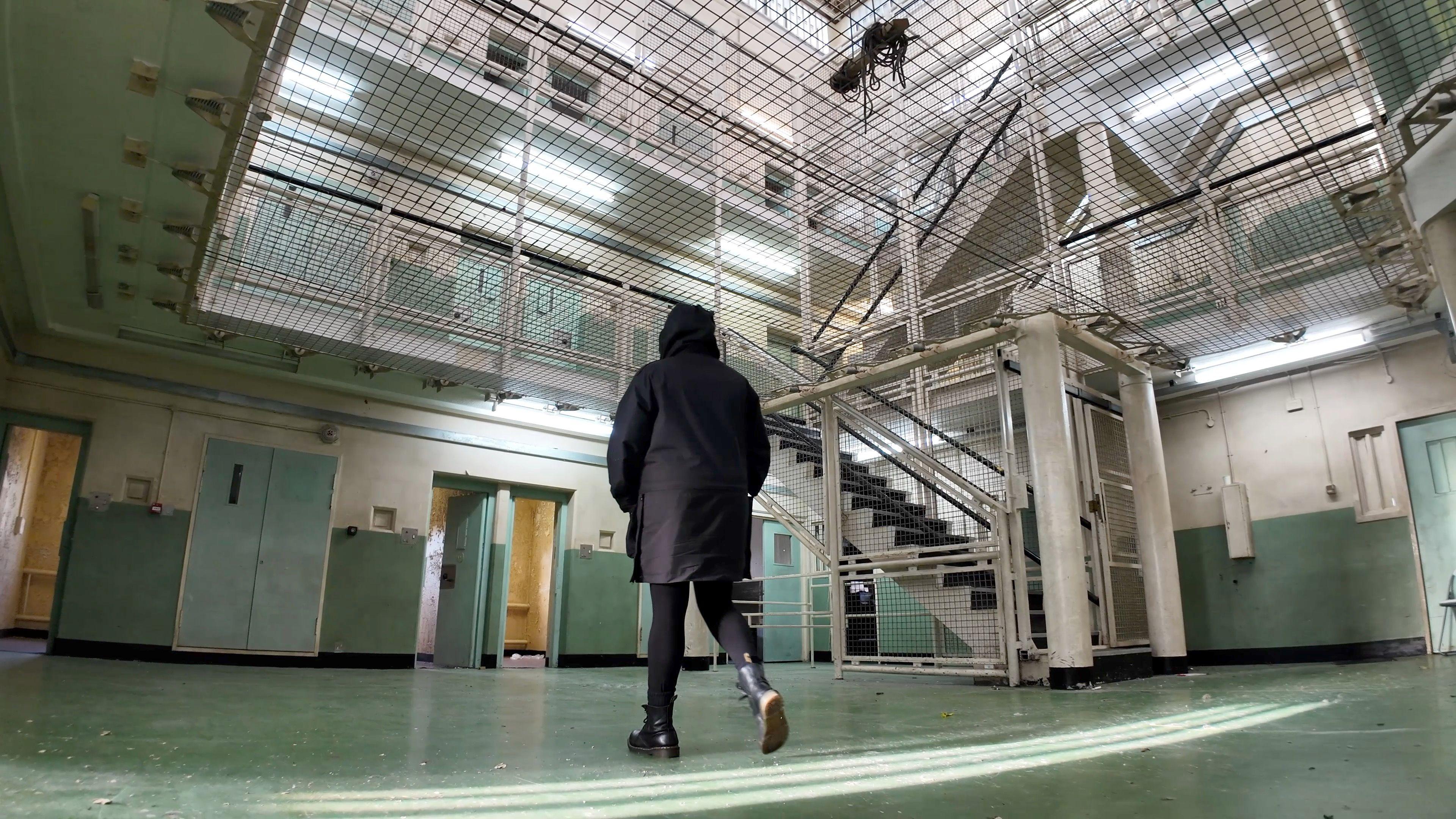 A former prison worker walking the landings of a former prison in Kent. He is wearing and dark hooded coat and is viewed from behind. The prison has a green floor, is stark and empty, with metal staircases and thick doors.