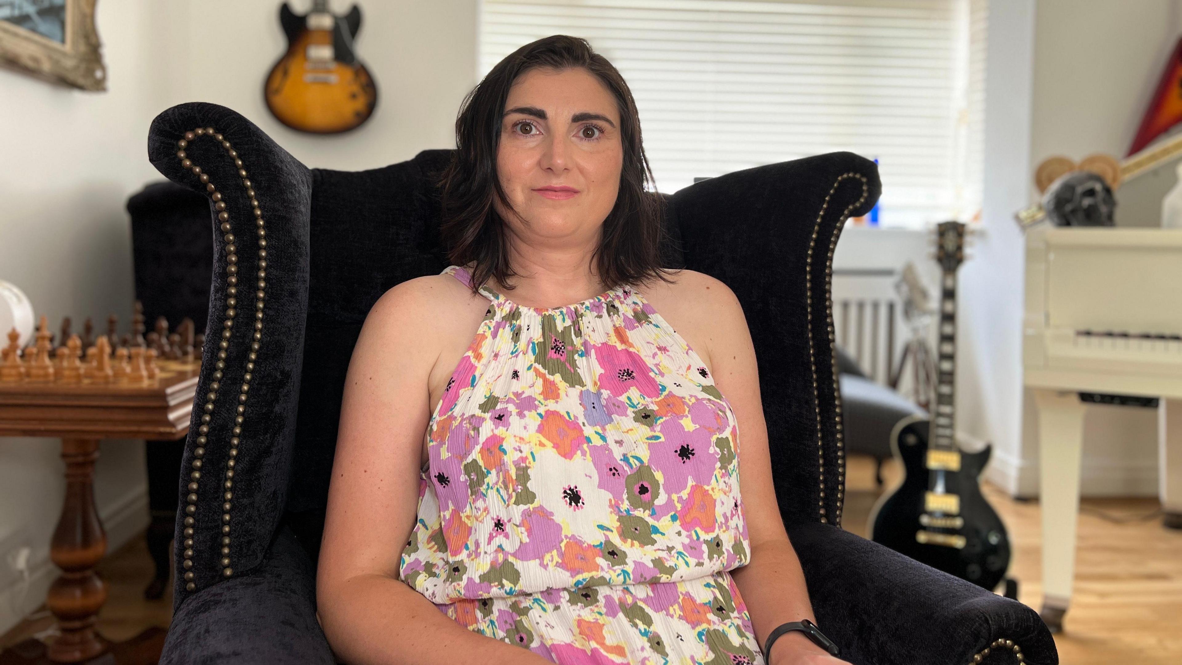 Dora sits in a black chair wearing a floral dress as she looks at the camera