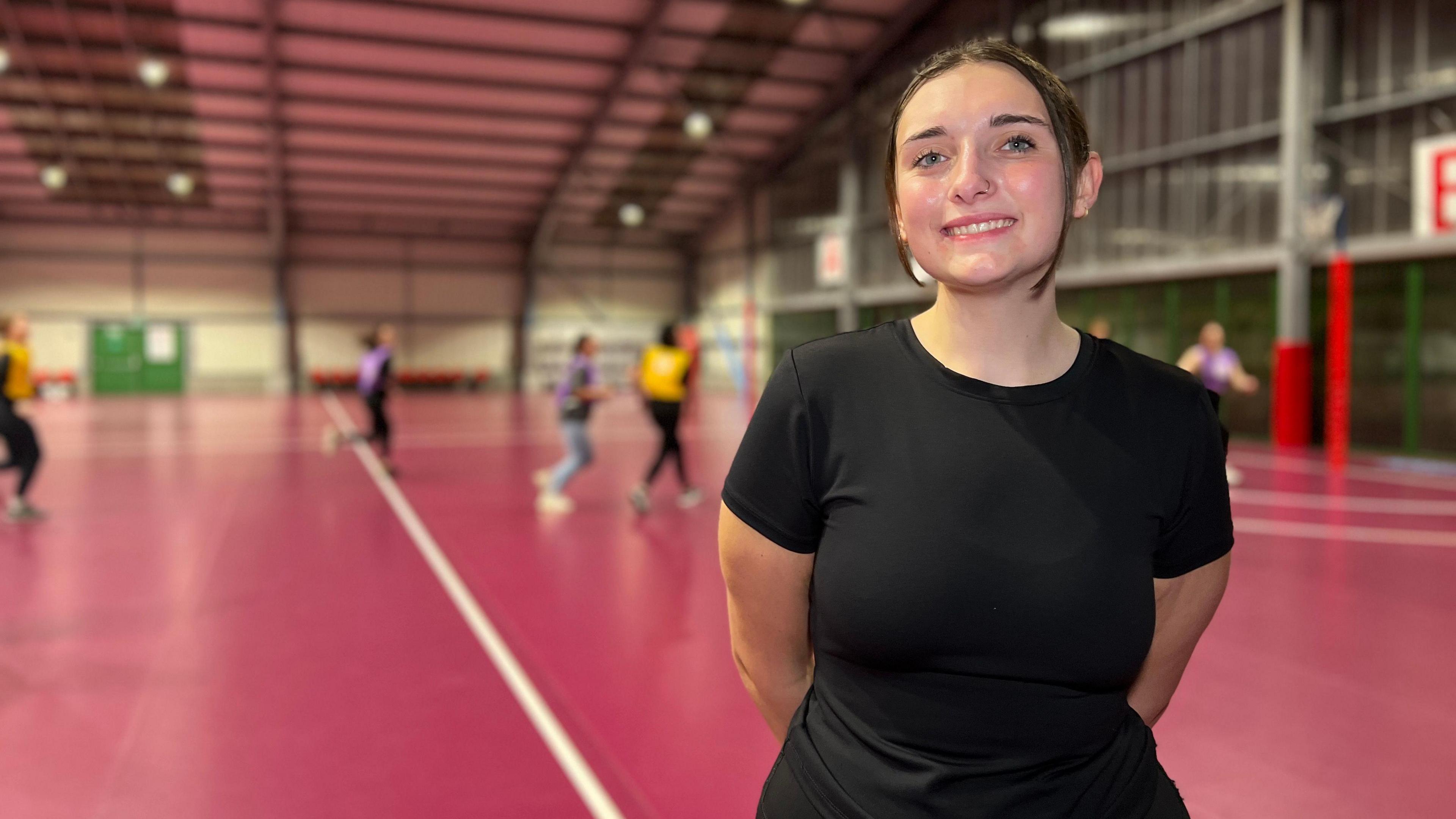 Kayla smiles at the camera and has her hands behind her back. She's wearing a black exercise top and her brown hair is tied back into a ponytail. Behind her people are playing netball on an indoor court.