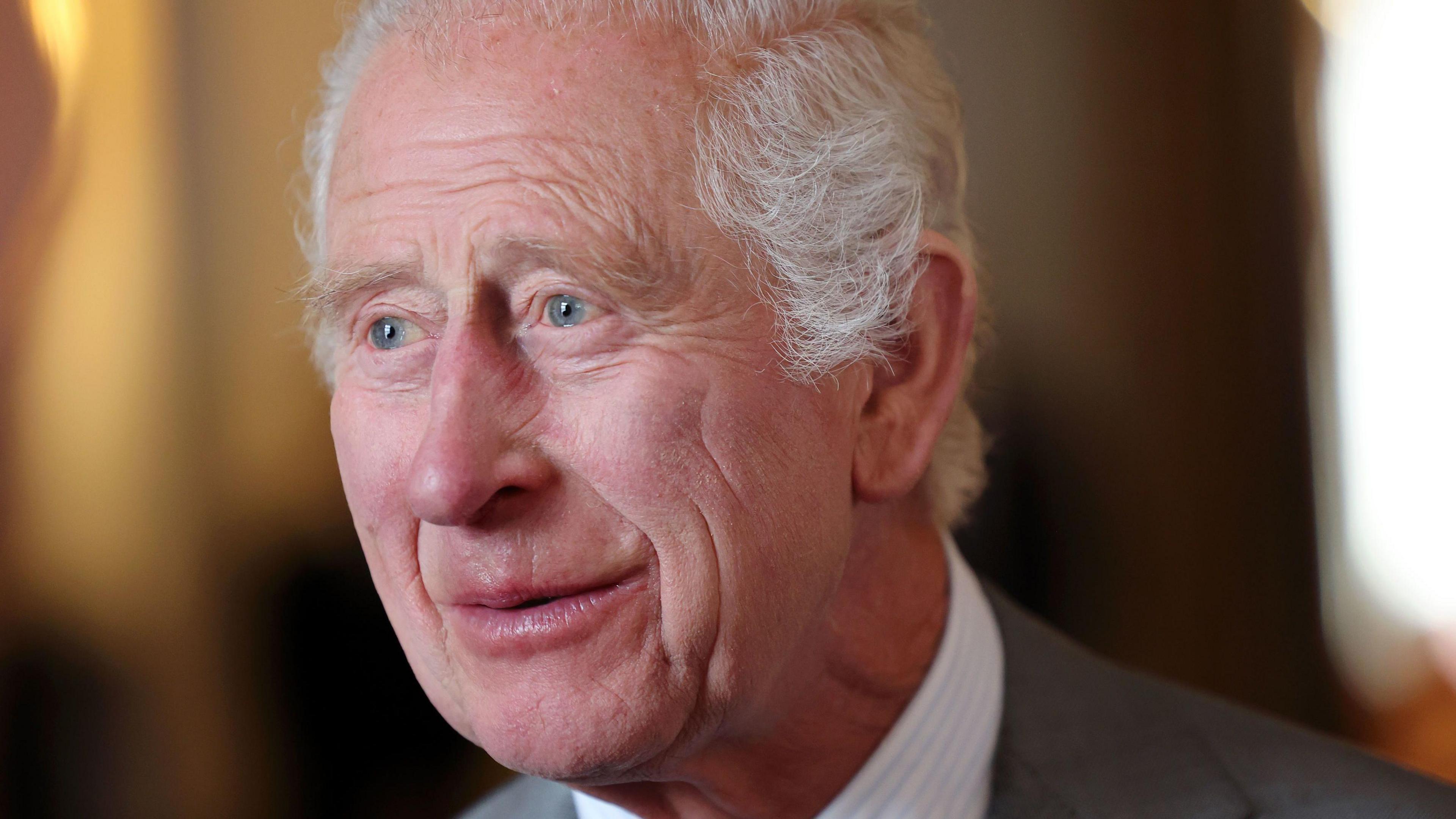 A close-up image of King Charles III's head. He is smiling to someone off camera. He has white hair and is wearing a grey suit jacket and a white shirt.