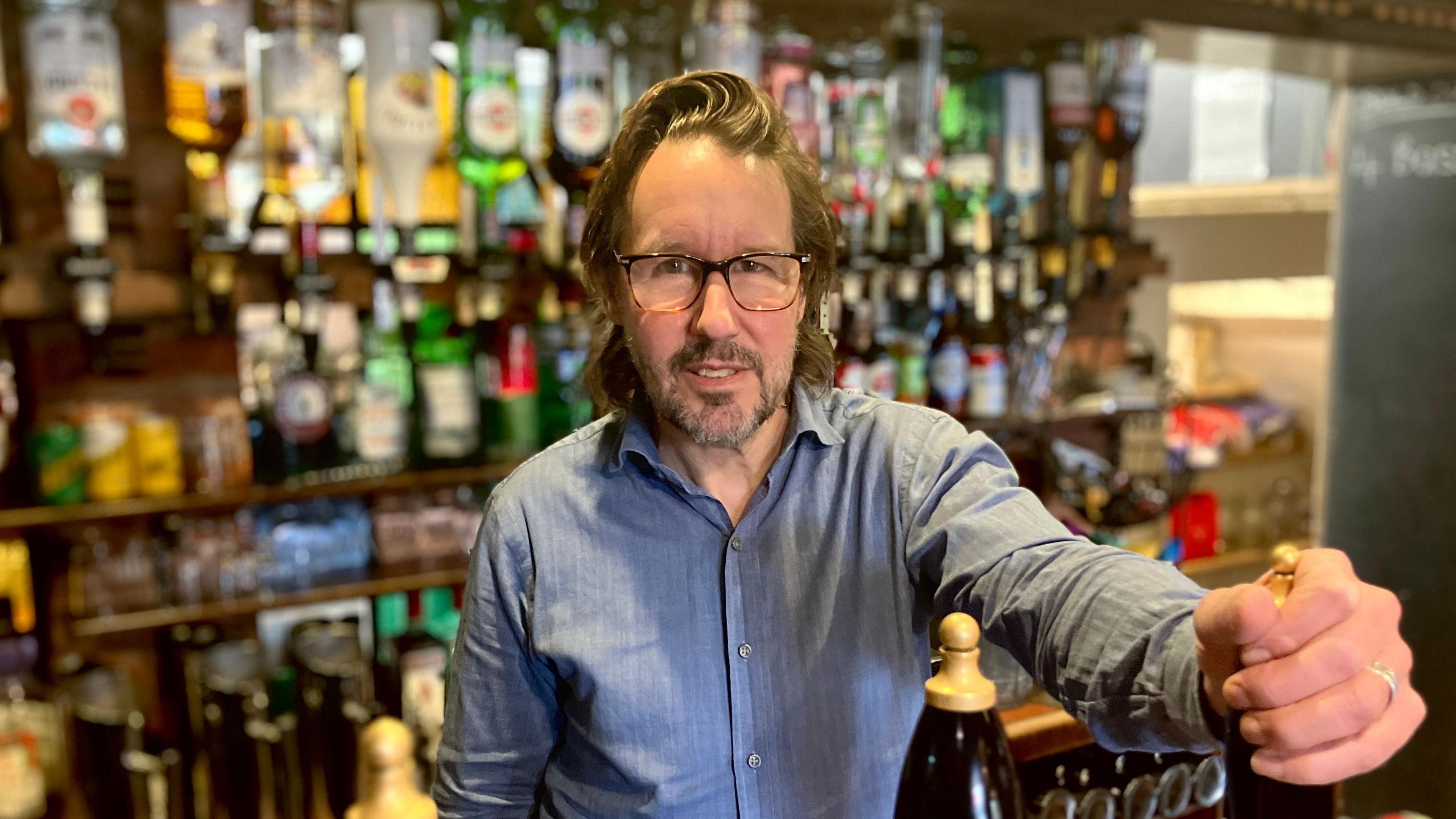 Venue owner Stuart Phillips standing behind his bar wearing a blue shirt with one hand on a beer hand-pump