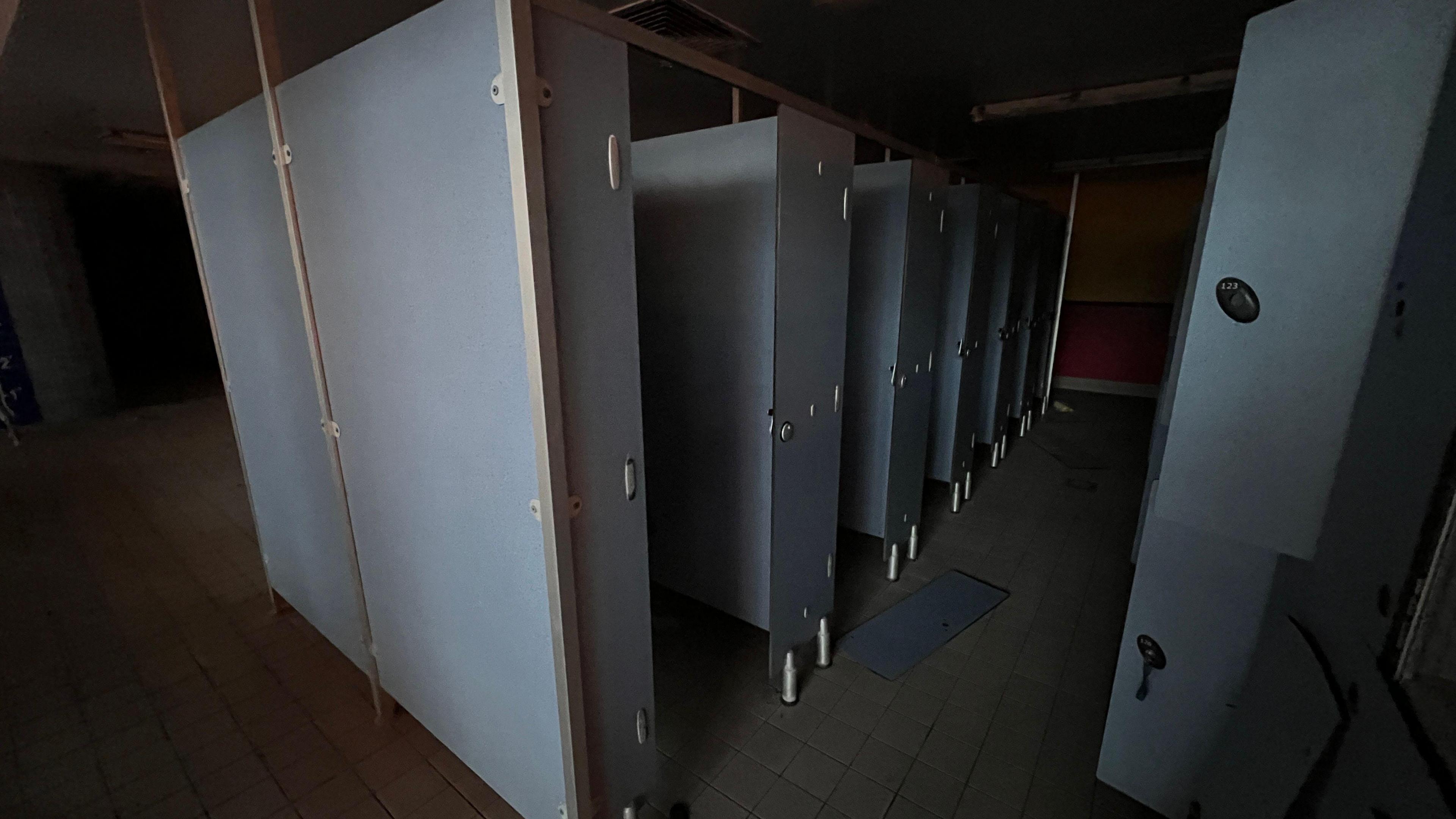 Image shows the changing stalls which are light blue and grey in colour. Five stalls are visible with the doors ajar and a white tiled floor below. Lockers are pictured to the right, with doors open, and are the same colour as the stalls. A broken locker door is visible on the floor in the centre of the image.