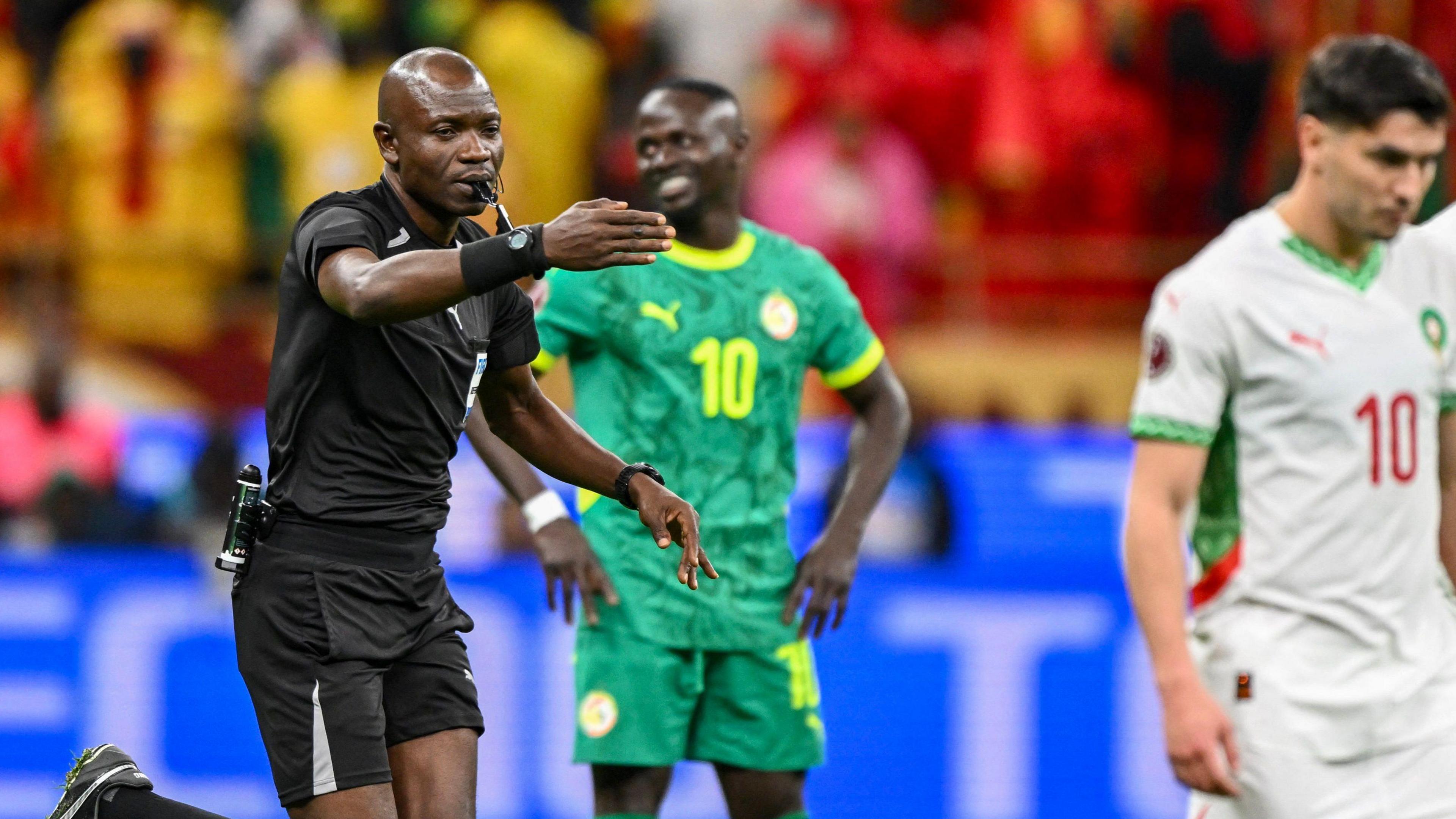 Referee Jean-Jacques Ndala points his right arm forward as he awards a penalty in the Africa Cup of Nations final. Out of focus in the background Sadio Mane can be seen with a wry grin on his face in t