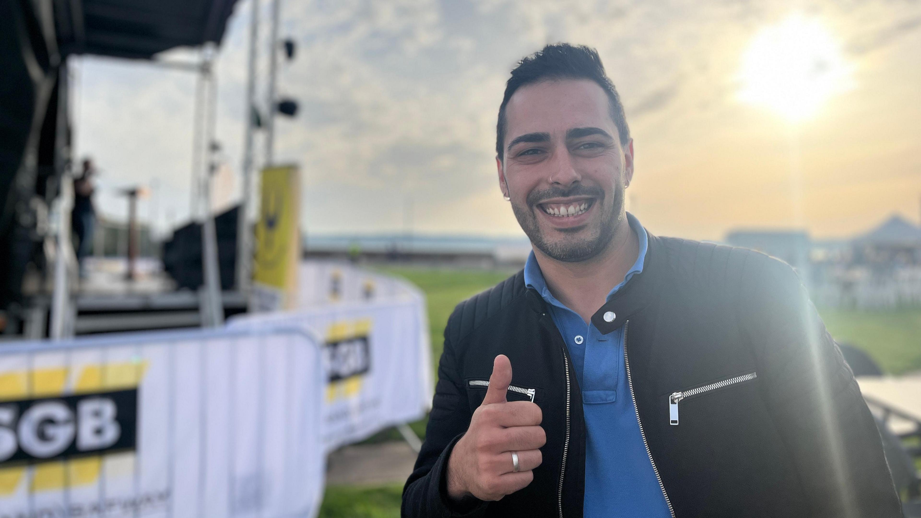 Telmo smiles at the camera as he gives a thumbs up. He has a black jacket on with a blue polo t-shirt. Behind him is the music stage and the sun is shining down on the field.