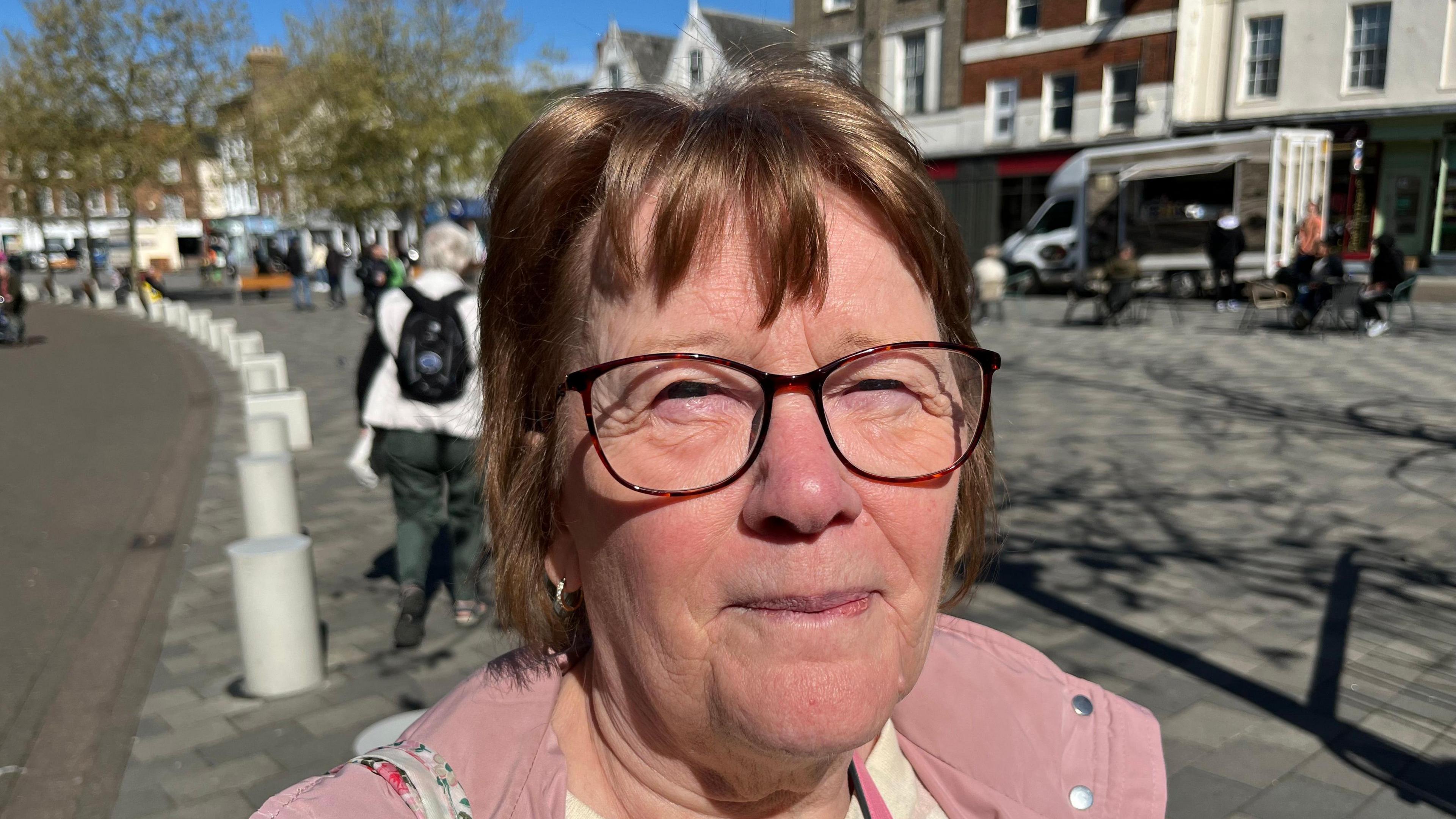 Janice Dunsdon, 75, has dark brown hair with brown thin rimmed glasses she is wearing a pink coat and behind her is the market place with shoppers and various buildings.