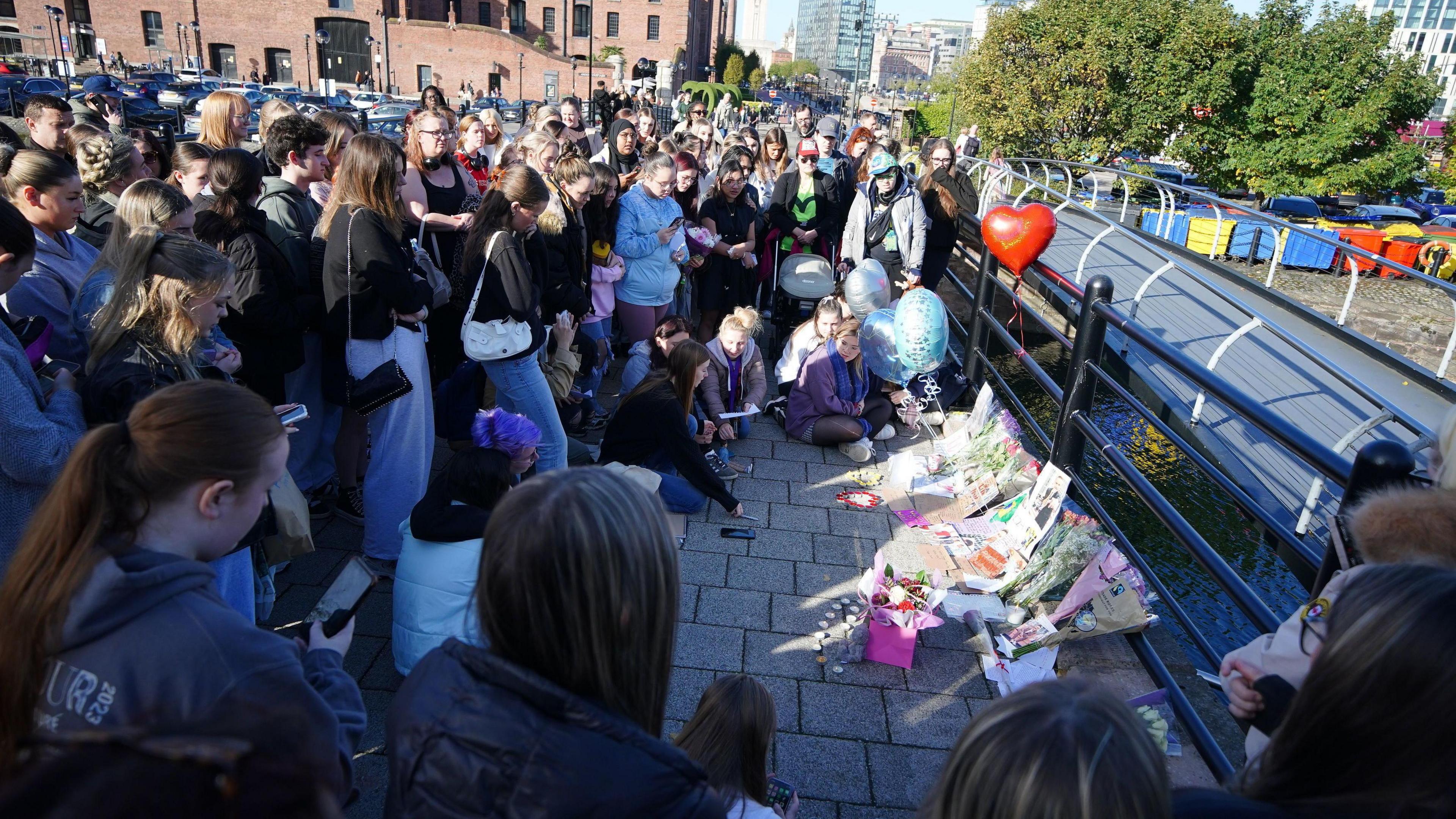 A crowd of about 50 people gather around a tribute consisting of bunches of flowers, balloons and notes leaning against railings on the dockside.