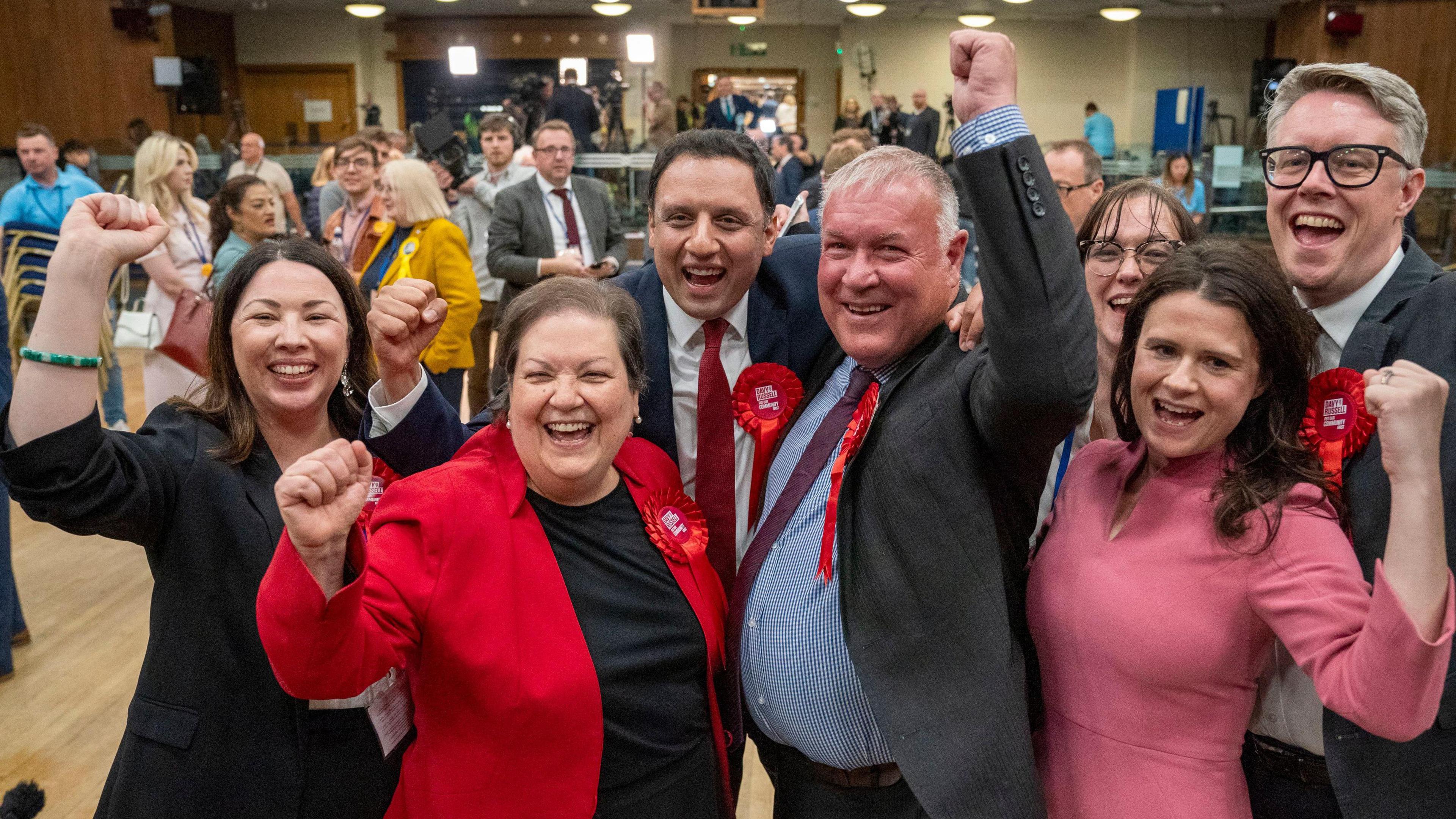 Labour politicians celebrating the party's victory in the Hamilton by-election, with leader Anas Sarwar - a man with dark hair and a red tie - and deputy Jackie Baillie - a shorter woman with dark hair and a red jacket - pictured at the count centre with candidate Davy Russell, a man with grey hair in a dark suit, wearing a red rosette, one clenched fist raised in triumph 