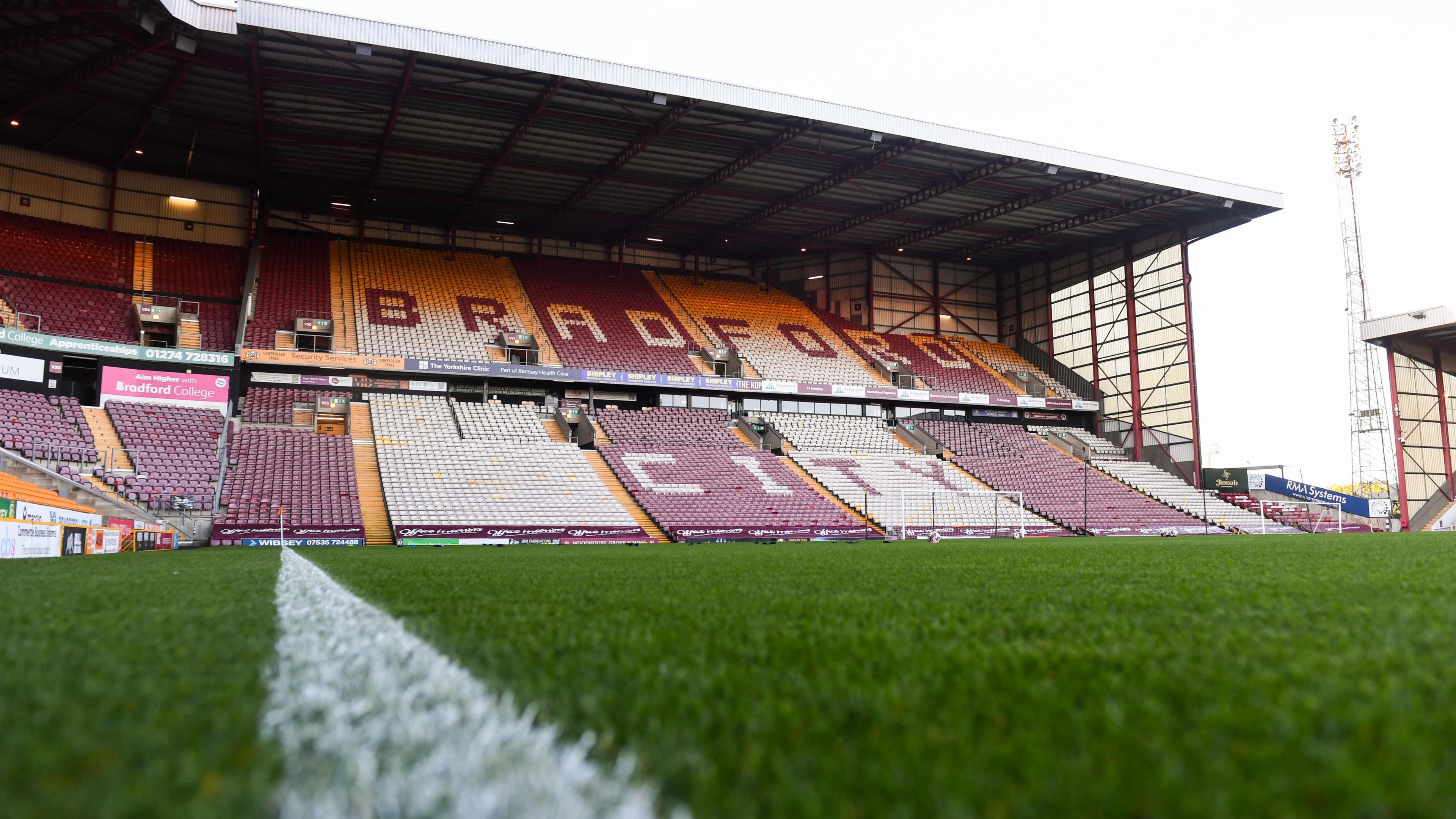 An empty football stadium, photographed from one of the pitch's touchlines. The words 'Bradford City' are visible across the seats in the stand visible.