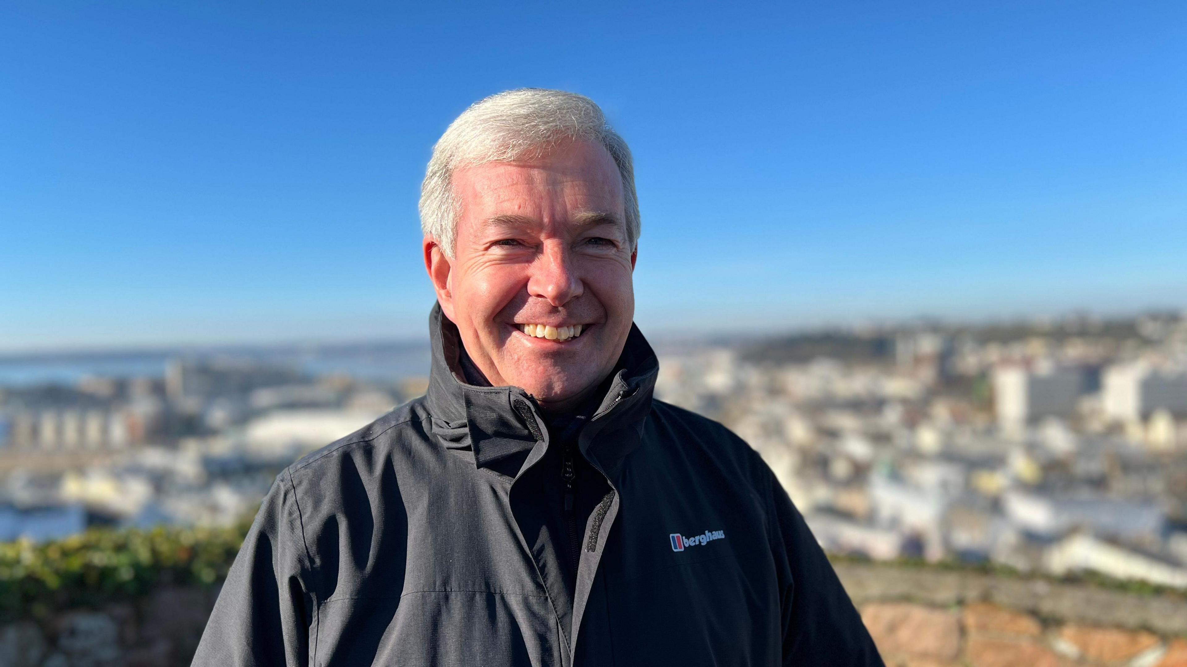 John Le Fondré is smiling at the camera, while standing on the ramparts at Fort Regent. An out-of-focus view of St Helier, with a blue sky, is behind him.