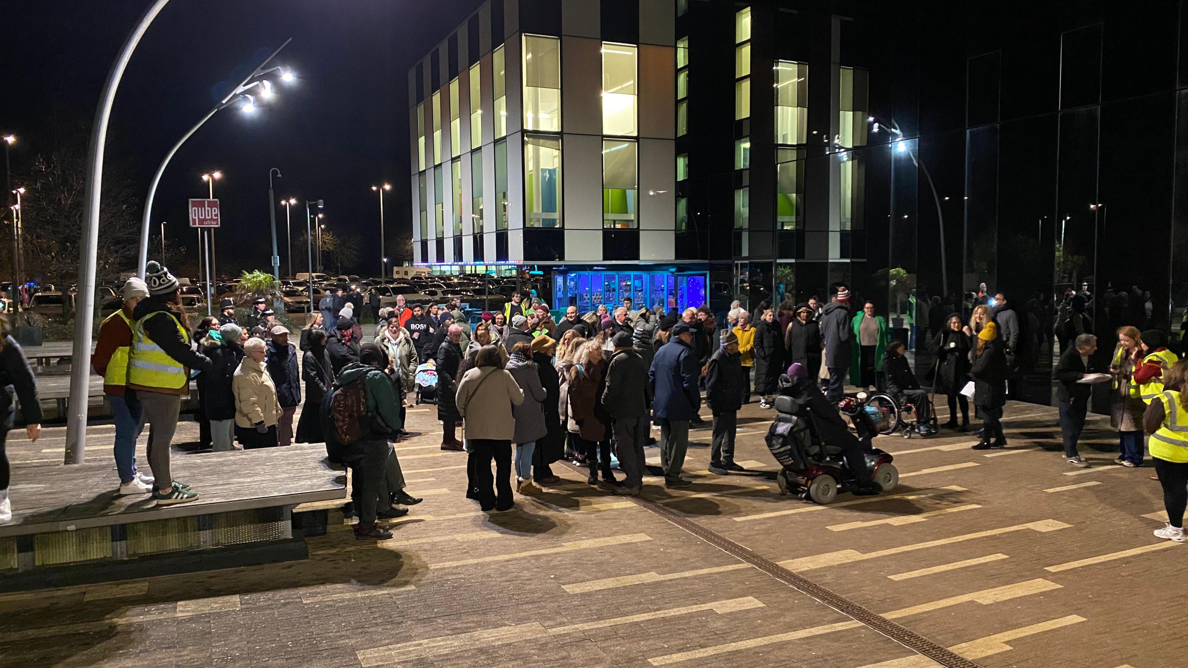 A crowd in coats and hats congregate outside the Corby Cube