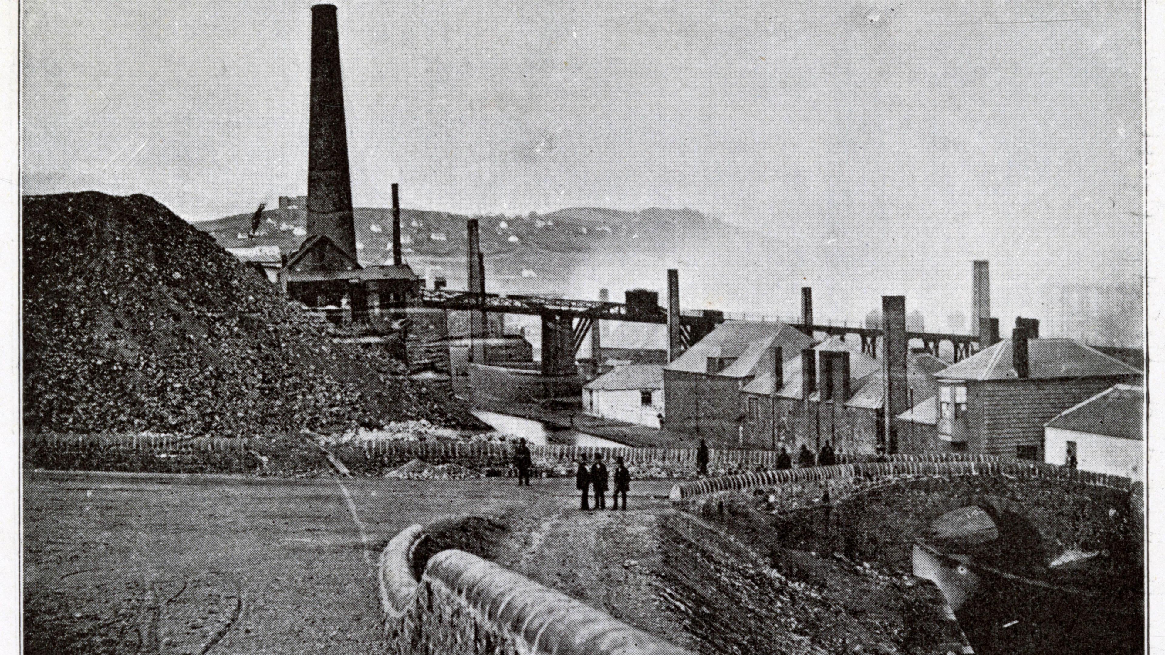 An historic image shows a range of historical industrial buildings at the copperworks site, with smoke seen in the background and about eight people seen standing near a little bridge over the river in the foreground.