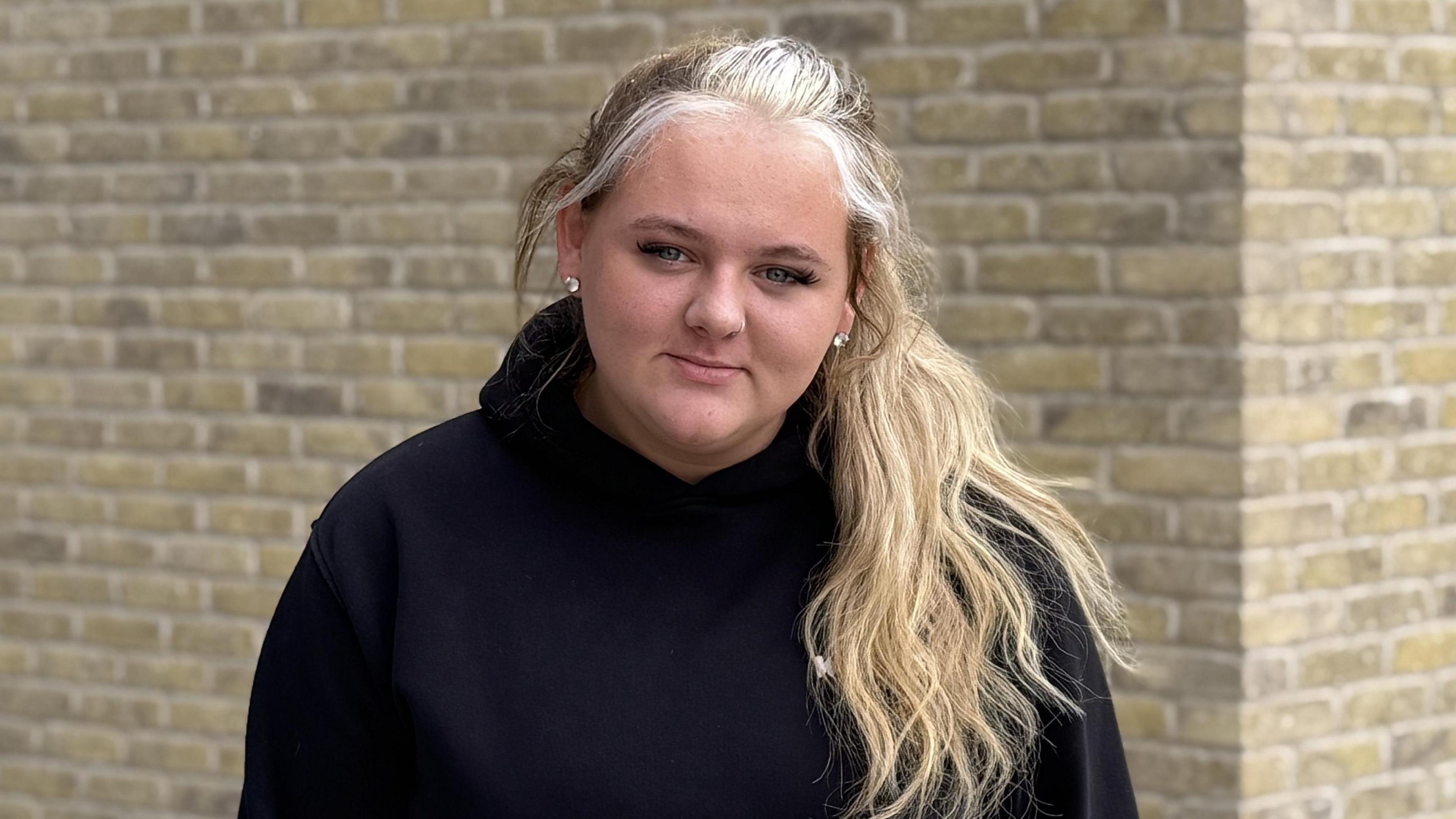 A woman with long blond hair is looking into the camera. She is wearing a navy blue hoody and is standing in front of a brown brick wall.