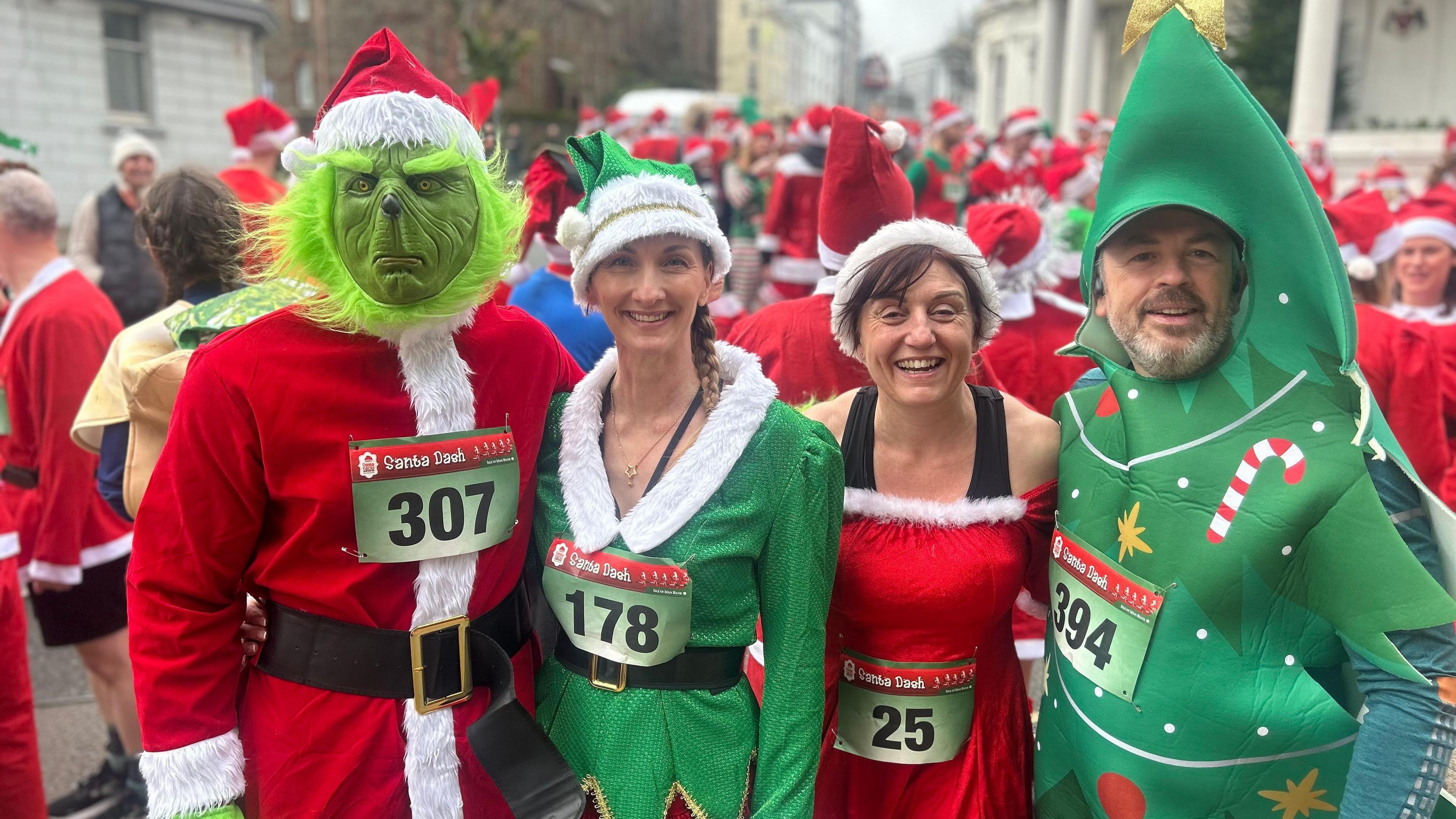 Runners smile for the camera dressed as the Grinch, a green character, an elf in a green suit, and a Christmas tree. A sea of santa hats can be seen in the background.