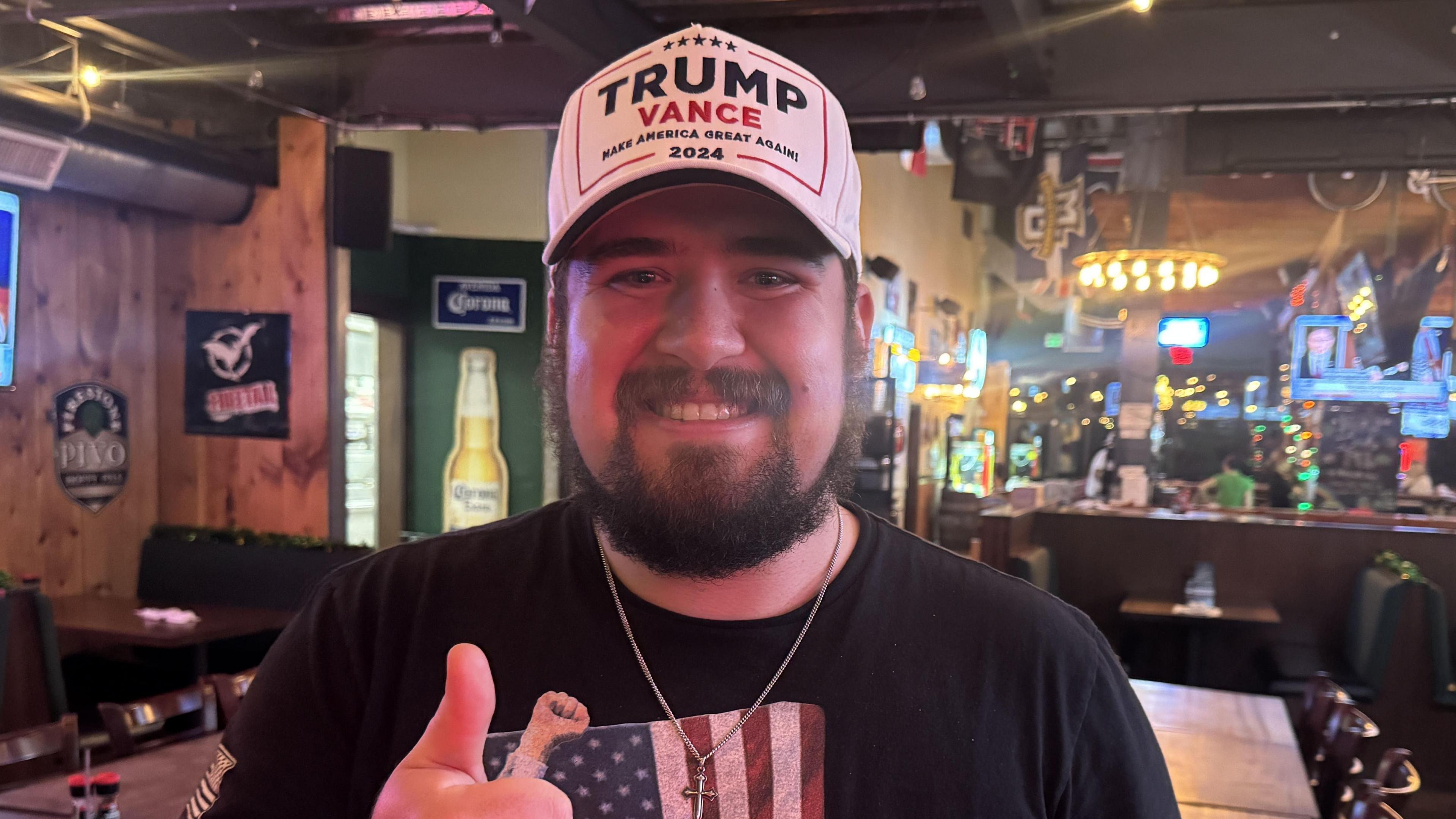 Evan Howard wearing a Trump Vance baseball cap, a US flag T-shirt and a crucifix necklace smiles at the camera with his thumb up