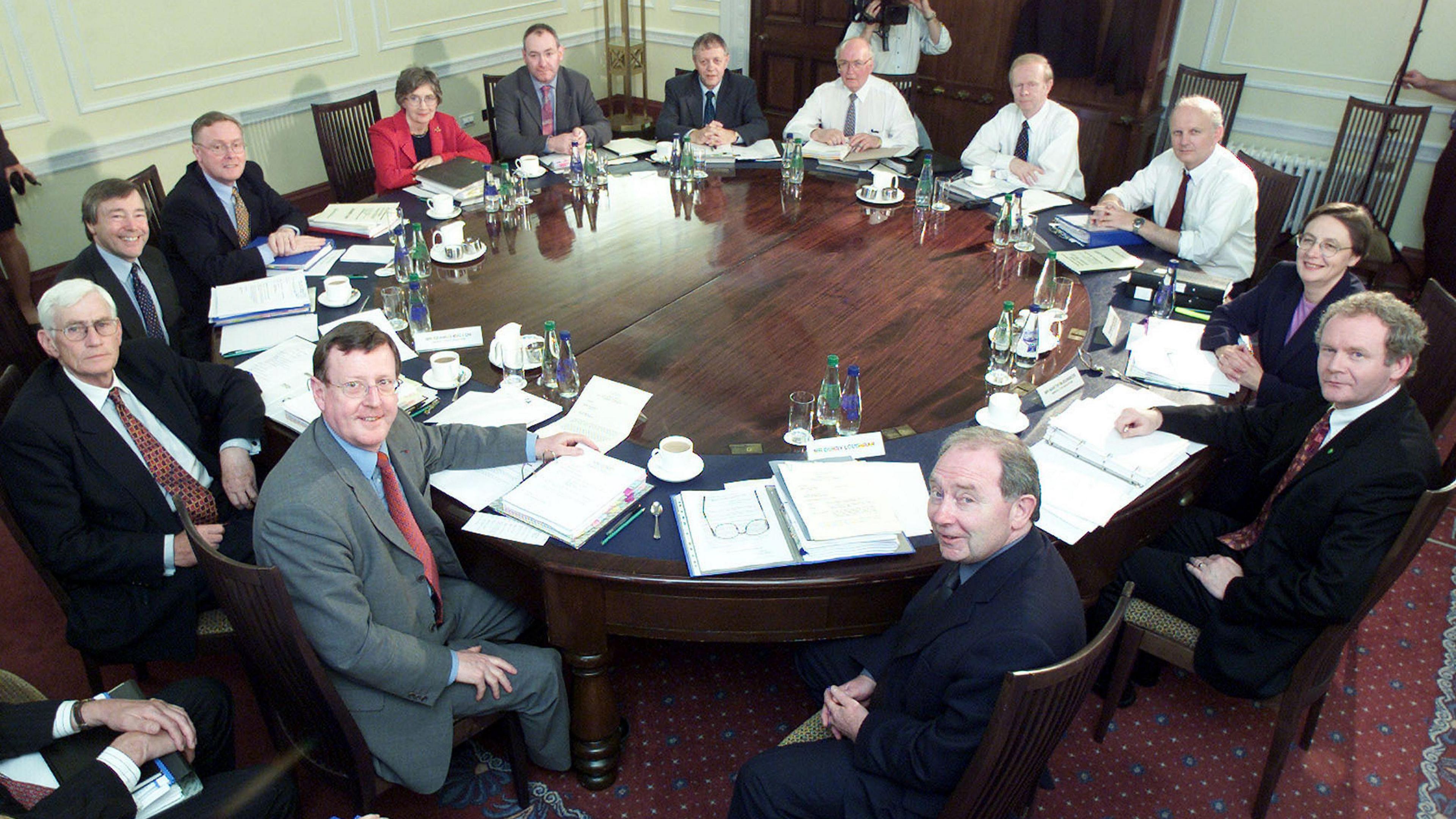 Northern Ireland Executive ministers and three civil servants seated at a round table at Stormont on 1June 2000.  Bríd Rodgers is sitting among them with her arms folded.  She is wearing a bright red suit jacket over a black top.