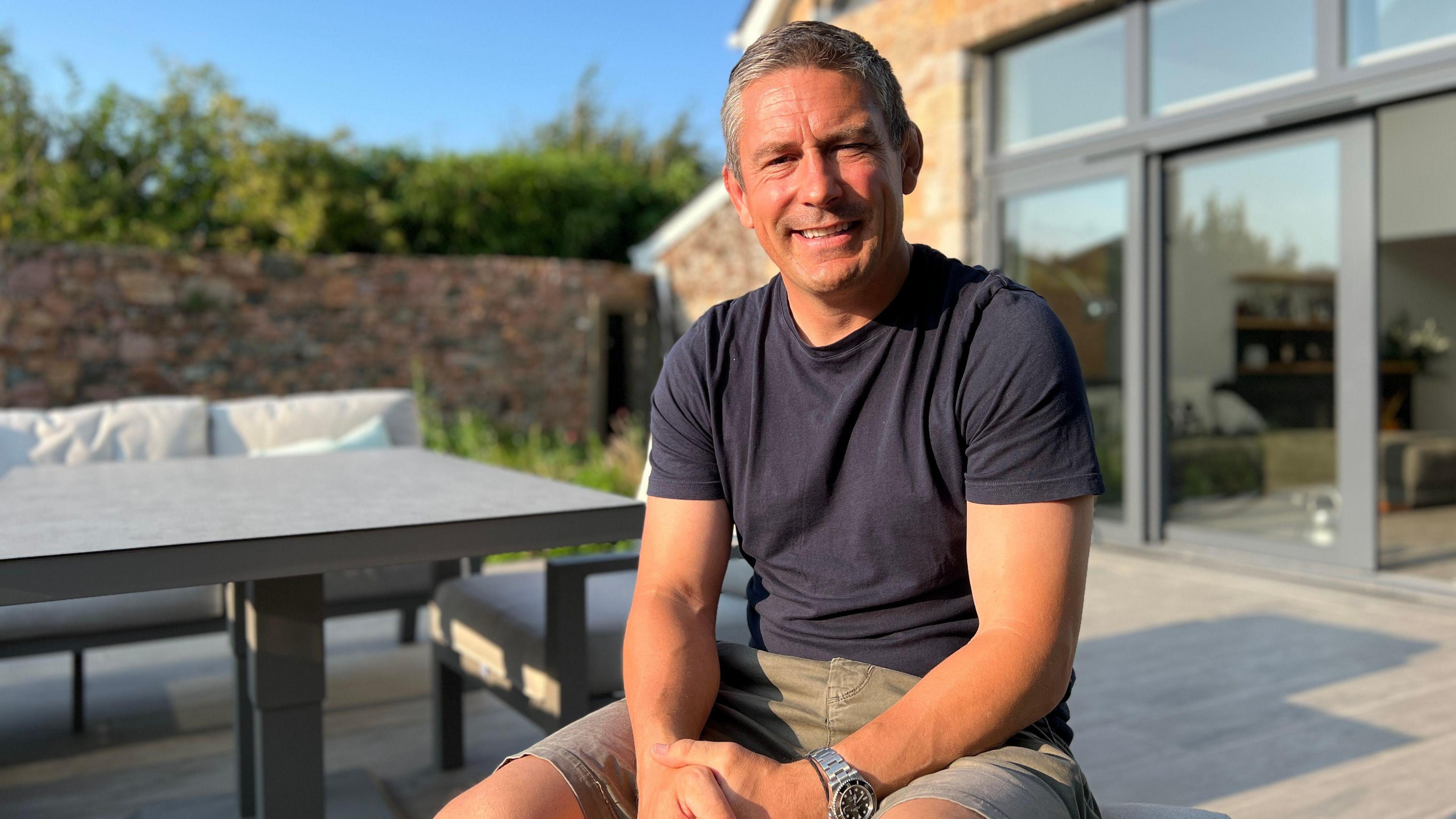 Graham smiles at the camera as he sits outside his house, next to a table with a garden wall and hedge in the background on a sunny day