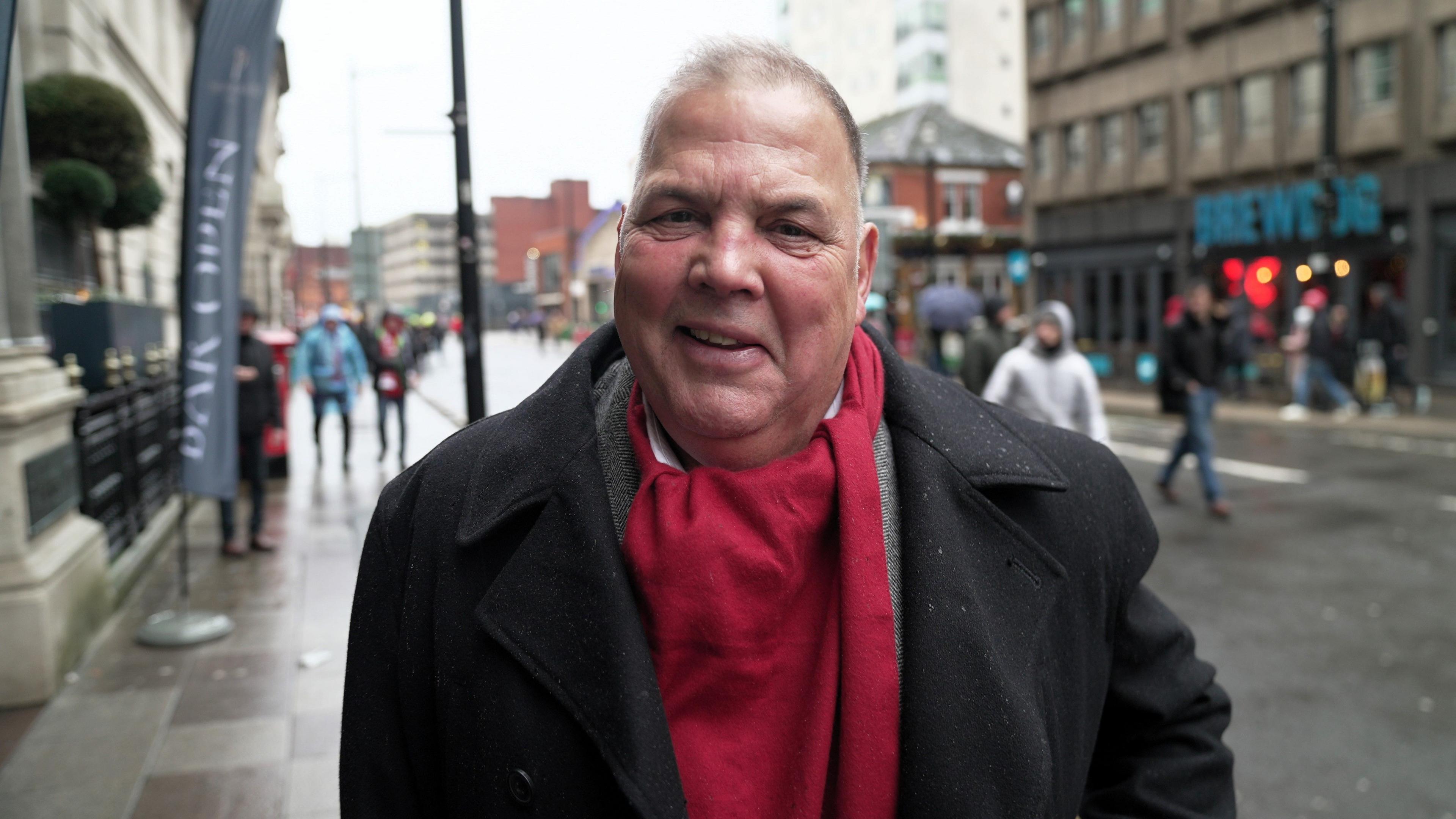 Nick Newman yn sefyll ar stryd yng nghanol Caerdydd, yn gwisgo cot ddu drwchus gyda sgarff coch llachar wedi’i lapio o amgylch ei wddf. Mae’r cefndir yn dangos adeiladau aml-lawr, siopau, a phobl yn cerdded ar hyd y palmant. Mae’r palmant yn edrych yn wlyb.