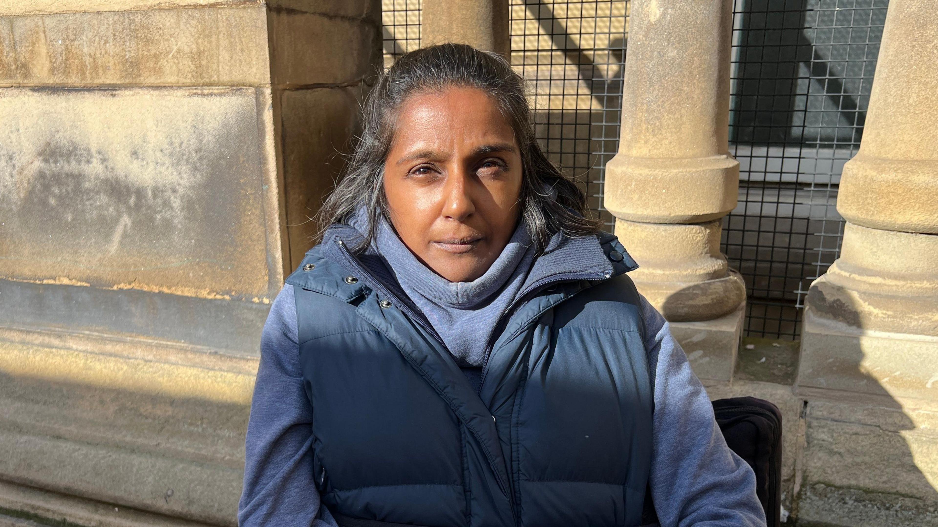 Rita Patel wears a navy blue jumper and navy gilet and is sat infront of the concrete of the council house in a wheelchair.