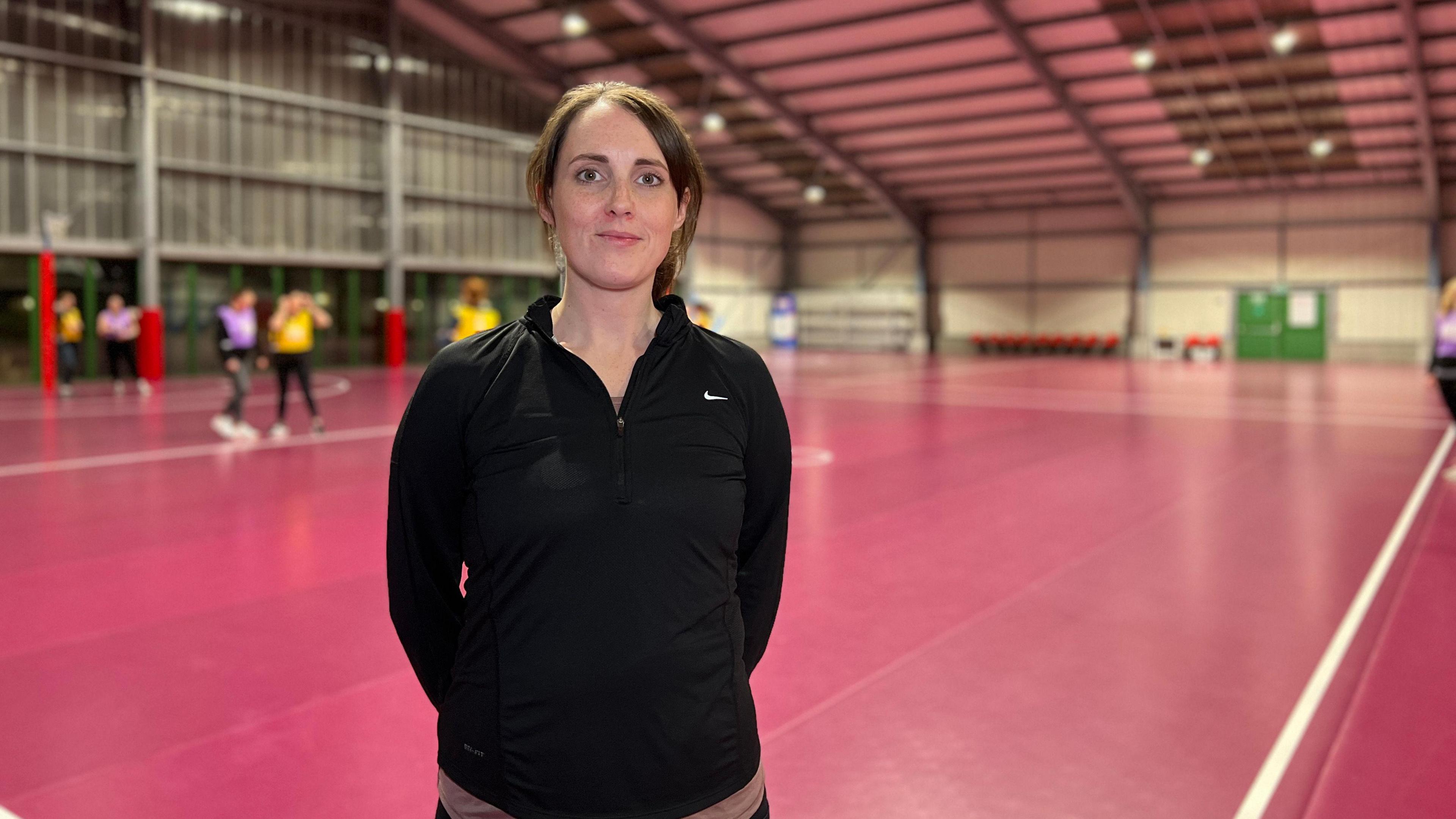 Sammy looks at the camera and her hands are behind her back. She's wearing a s black sports jacket and she has brown hair which is tied into a ponytail. Some people play netball on an indoor court behind her.