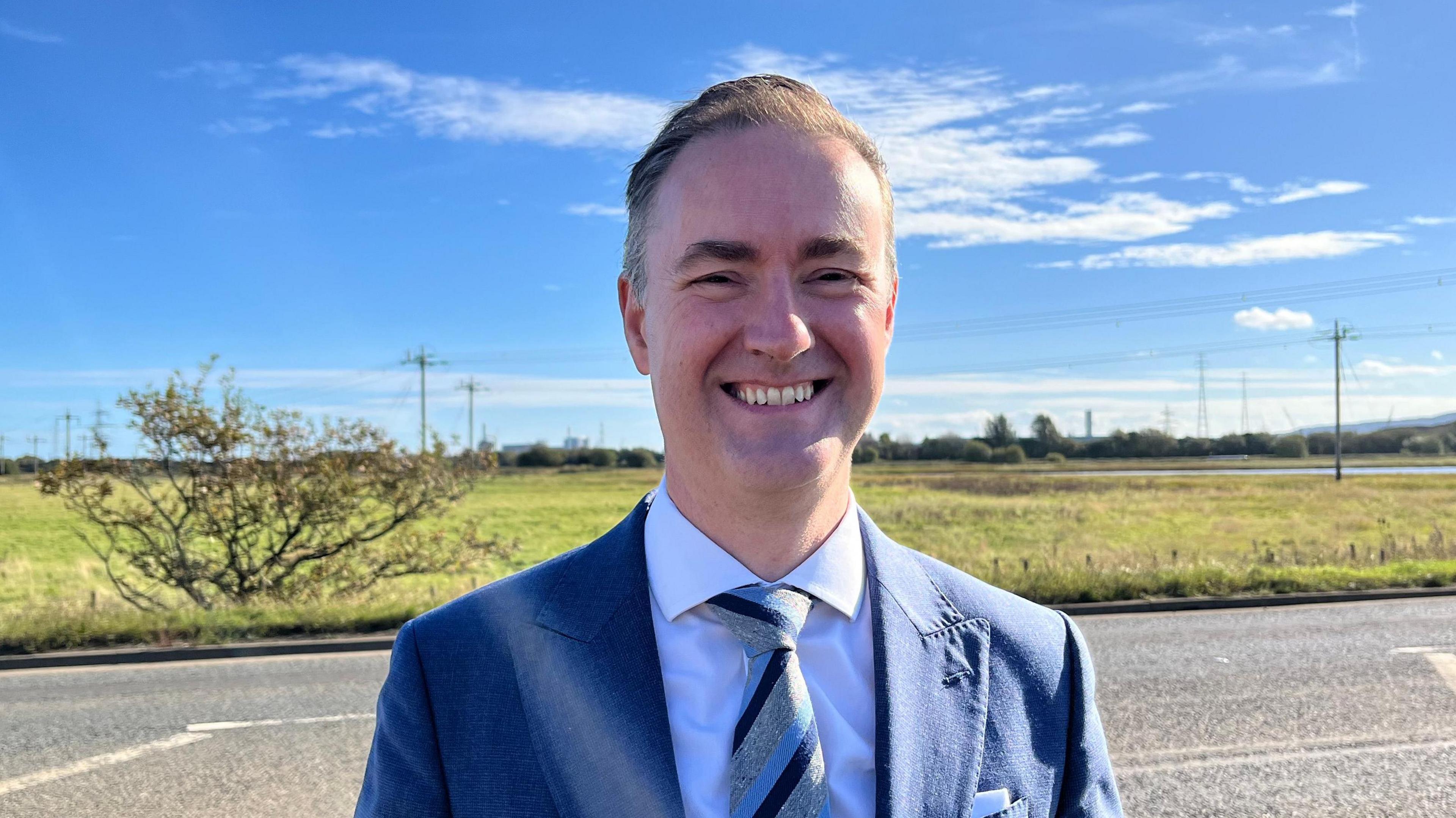 MP Chris McDonald is dressed in a blue suit and smiling. In the background there is a road and field with a bush to his left and power lines to the right.