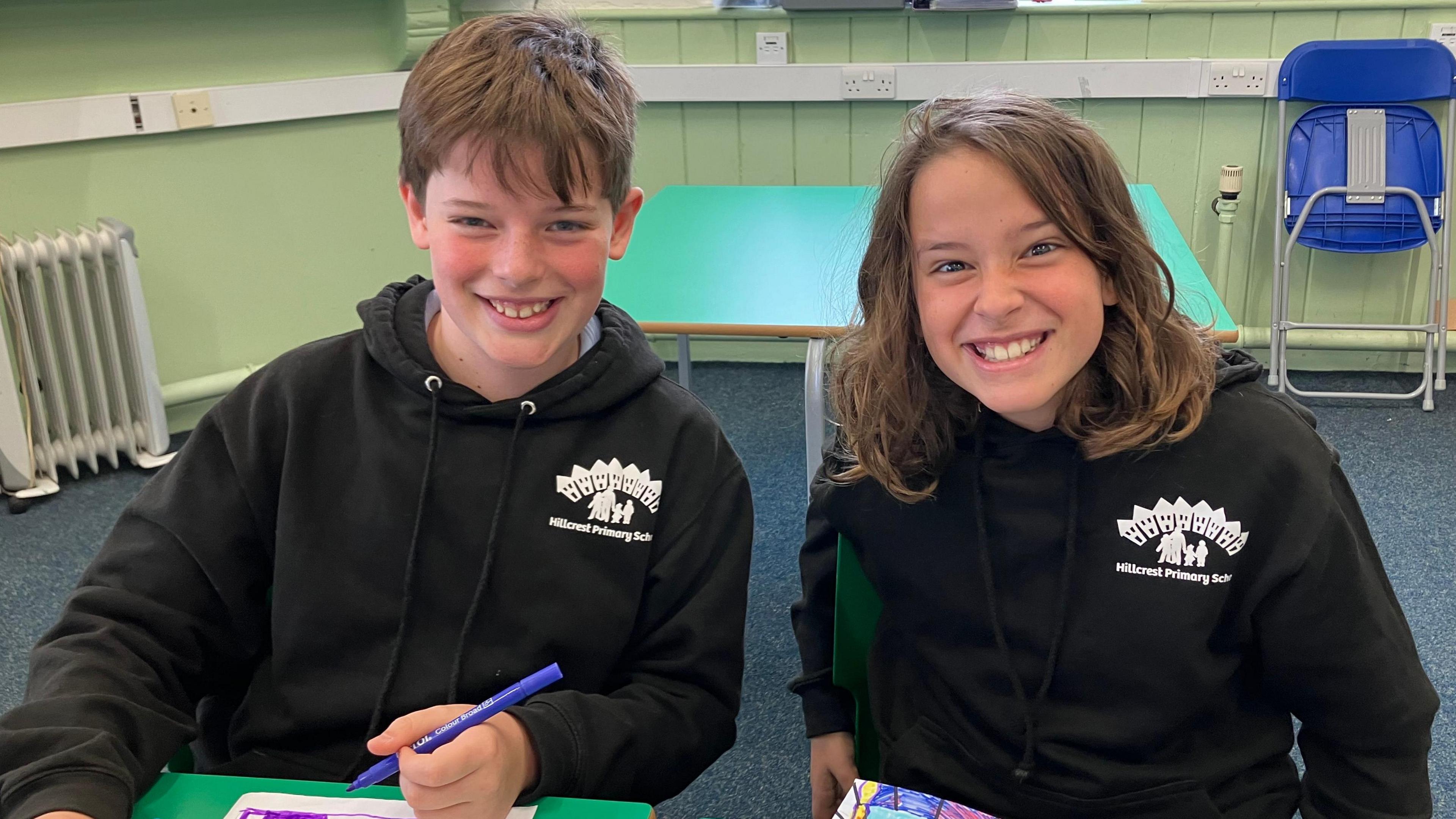 Twins Felix, who has short brown hair, and Rudy, who has long brown hair, sit together at a green table in class. They are wearing black school leavers hoodies and smiling